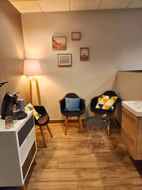 Coin salon cosy avec trois fauteuils, œuvres d'art, lampe et coin café. Couleurs chaudes, parquet.