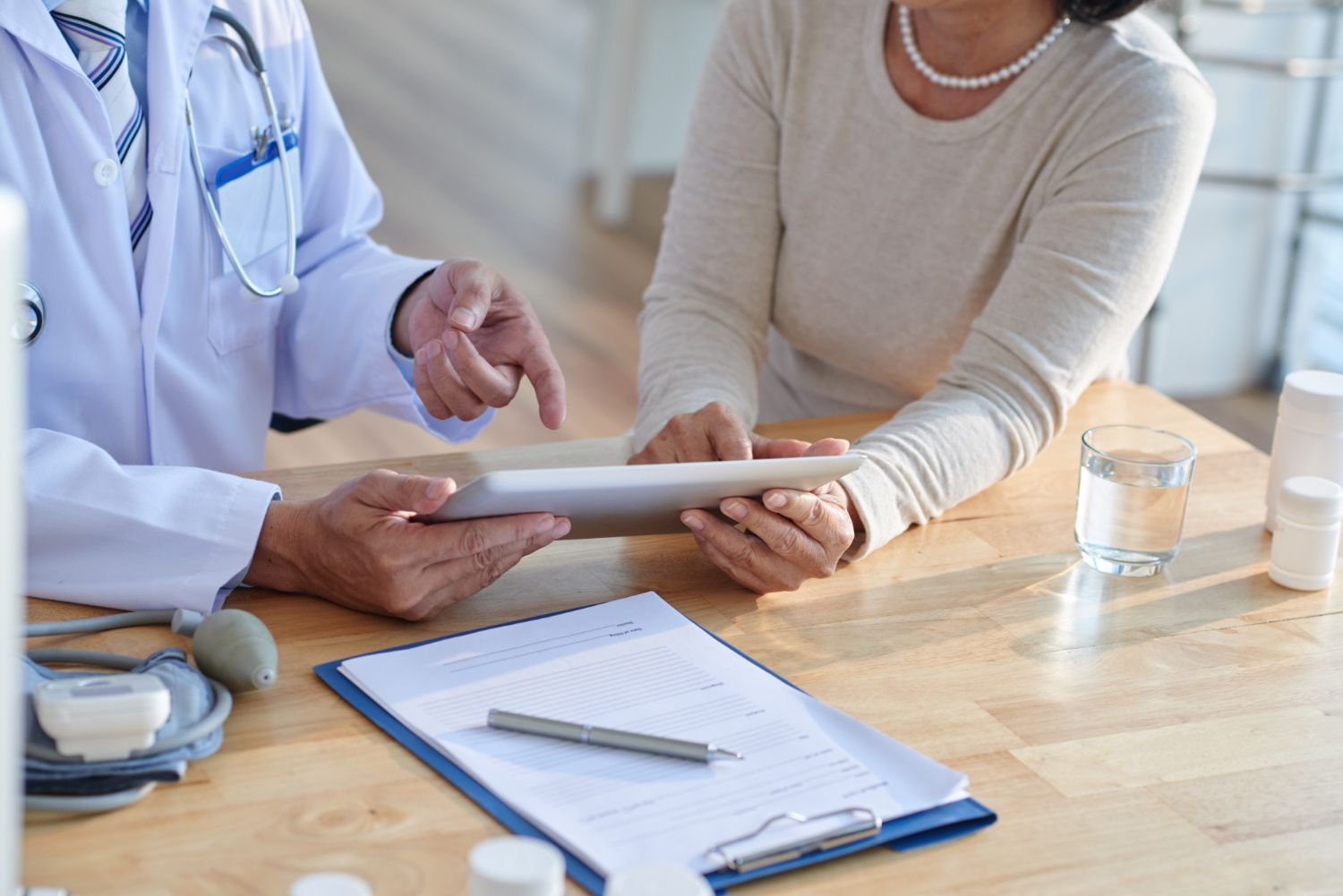 A healthcare provider in a white coat sits with a patient, reviewing information on a tablet at a table with medical forms.