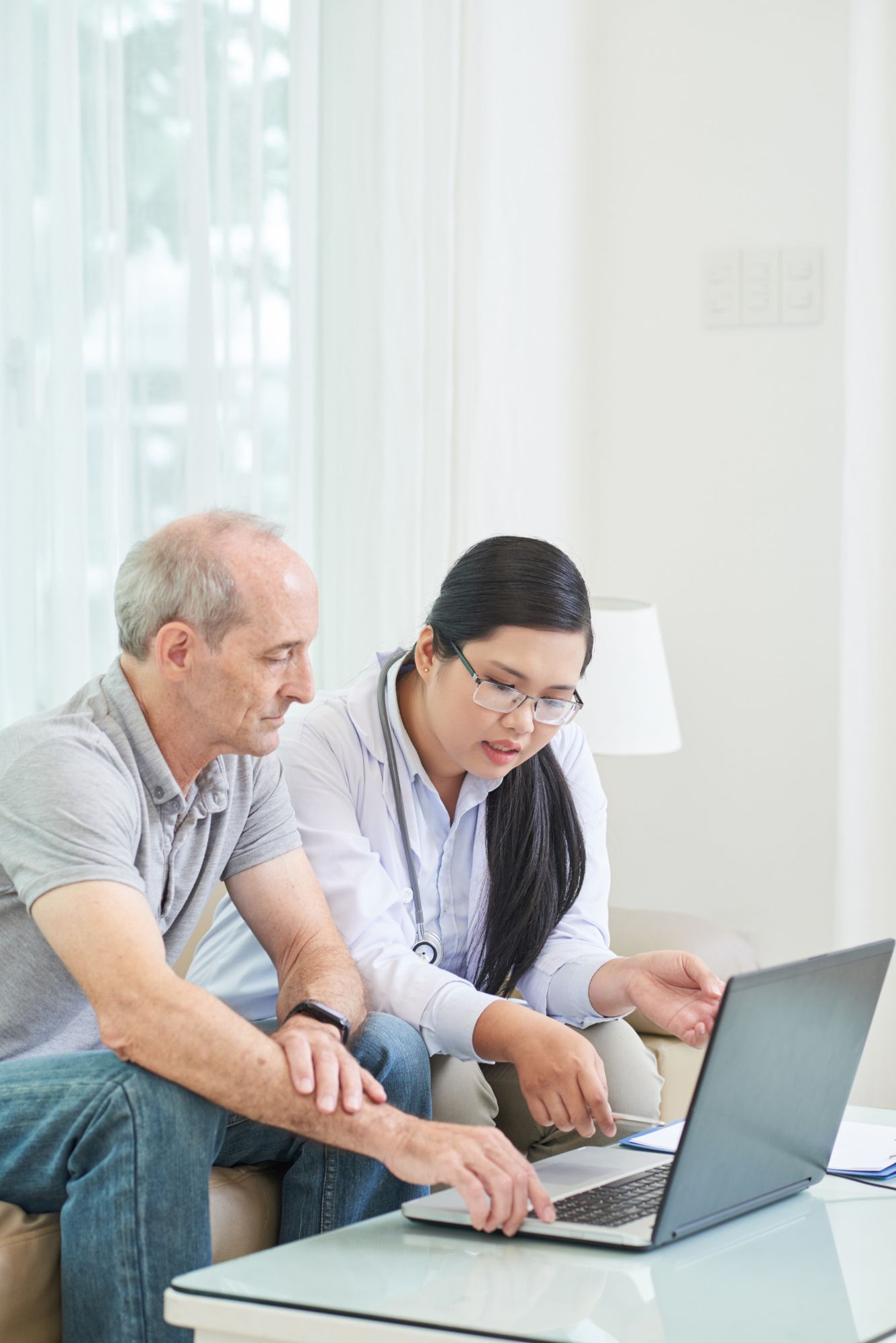 A healthcare professional wearing a stethoscope sits on a sofa, explaining information on a laptop to a patient.