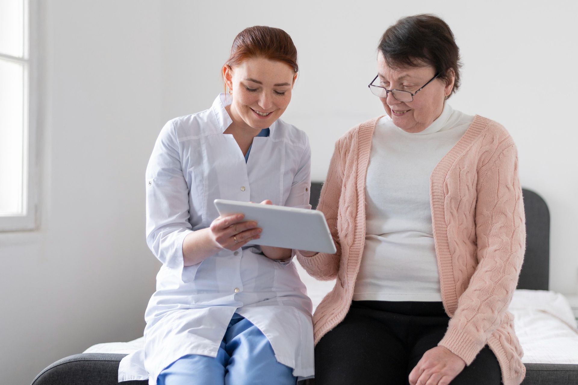 A healthcare professional in a white coat shows information on a tablet to a patient while sitting together on a bed.