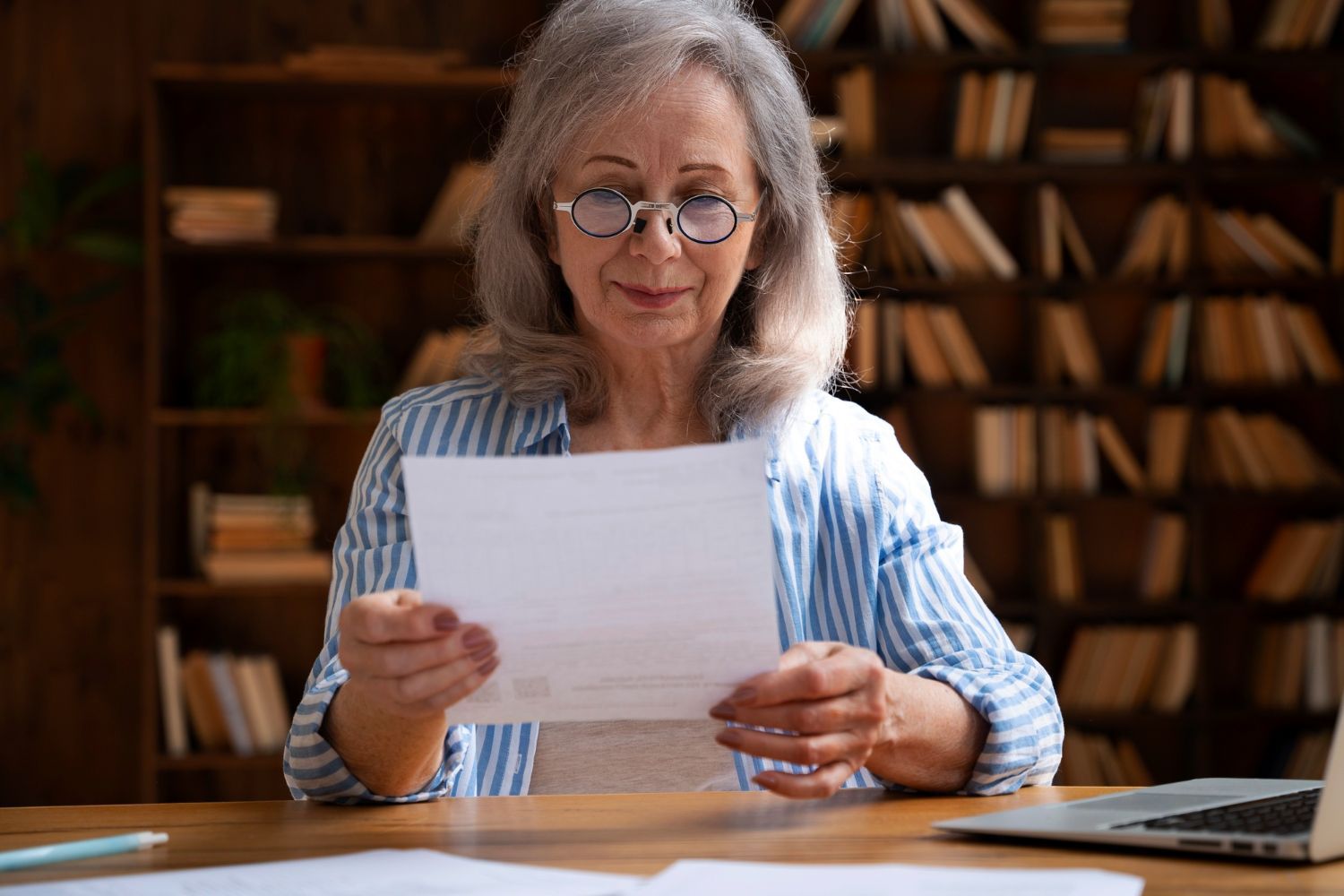 A person in a striped shirt reads a document at a desk in a library filled with bookshelves.