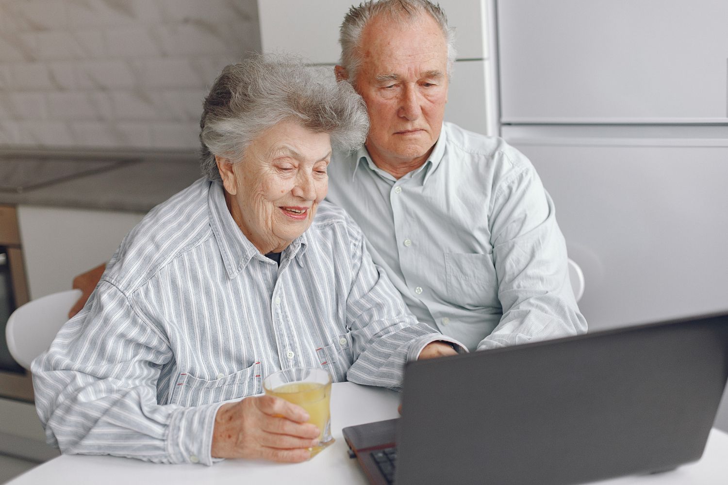 Two people seated at a table looking at a laptop, with one holding a glass of juice in a bright, modern kitchen.