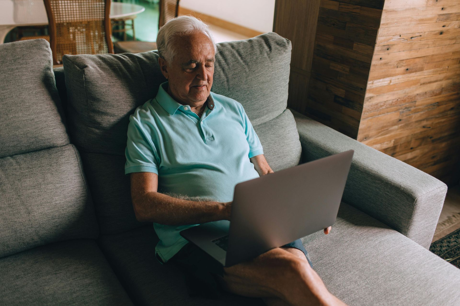 A person wearing a light blue polo shirt sits on a grey sofa, typing on a laptop.