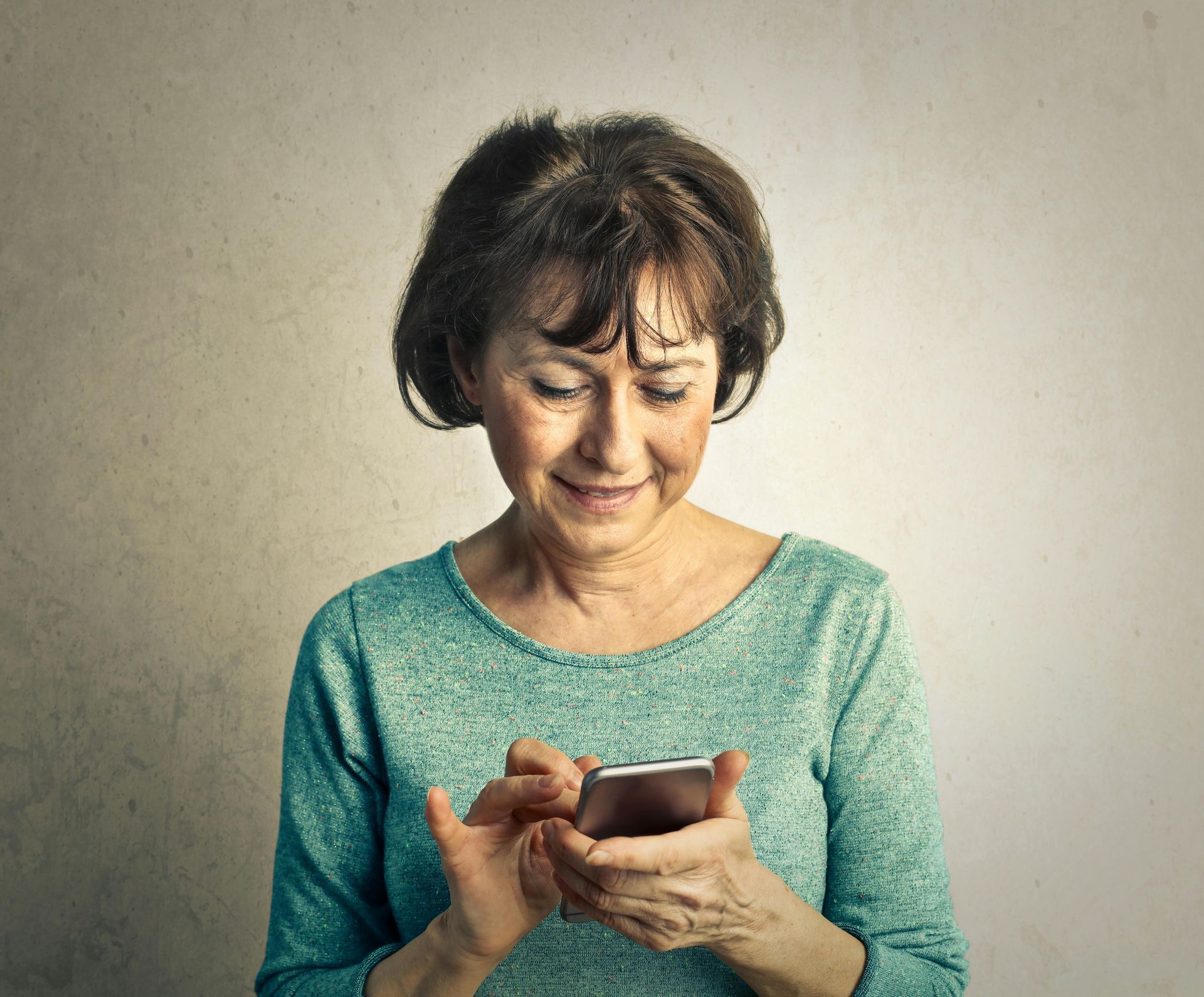 A person in a green long-sleeved shirt looks down and smiles while using a smartphone against a plain wall.