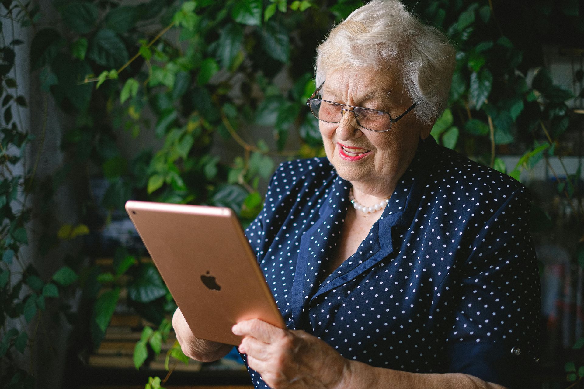 An older person wearing glasses and a blue polka-dot blouse smiles while using a tablet in front of a green leafy plant.