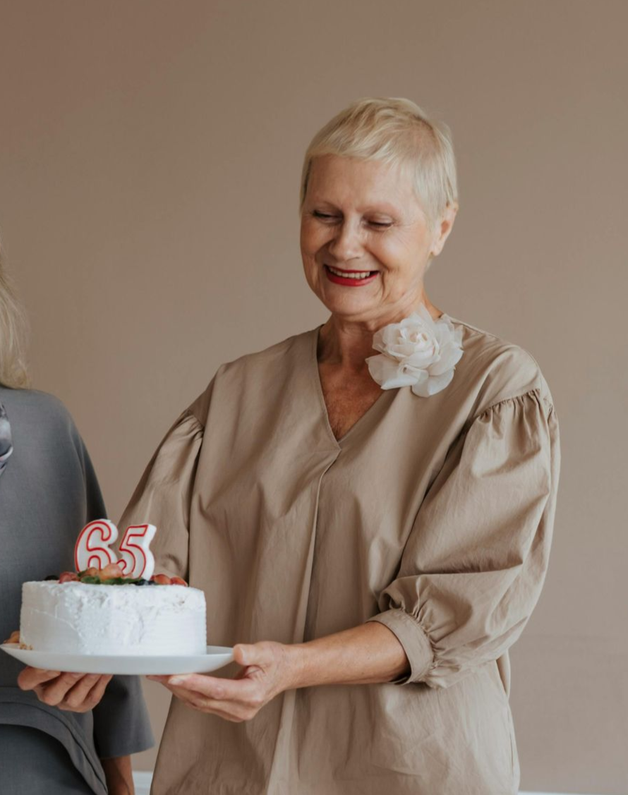 A person smiles while holding a white cake topped with a candle shaped like the number 65.