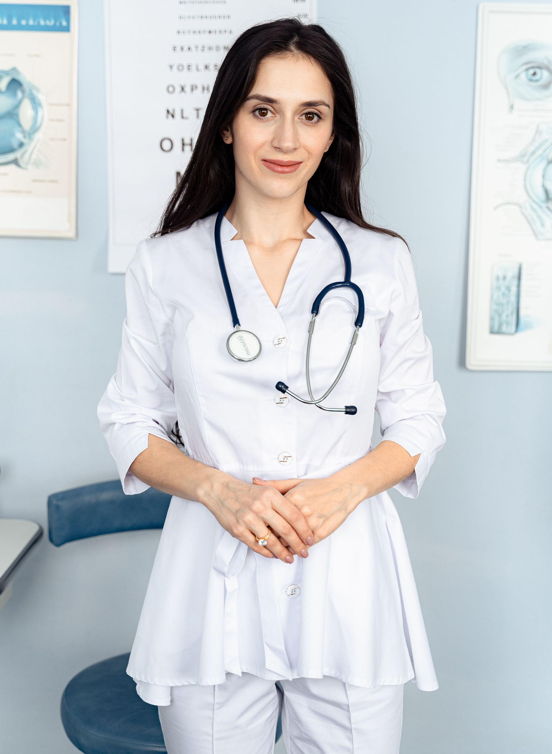 A smiling person in a white lab coat, stethoscope, and blue gloves holds a clipboard and a small pill bottle.