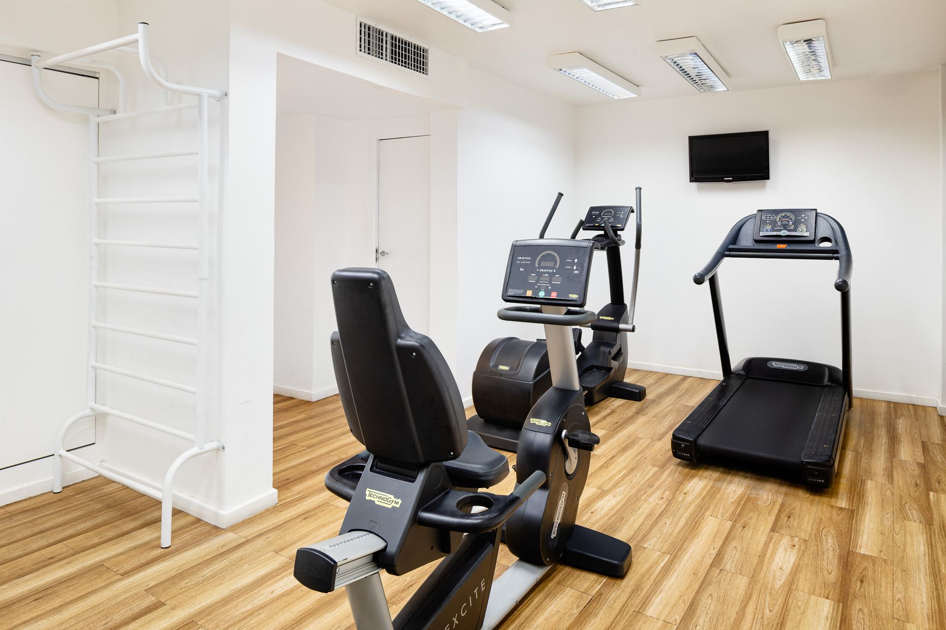 Academia com equipamentos de ginástica: bicicleta ergométrica reclinada, elíptico, esteira, TV e prateleiras contra uma parede branca e piso de madeira.