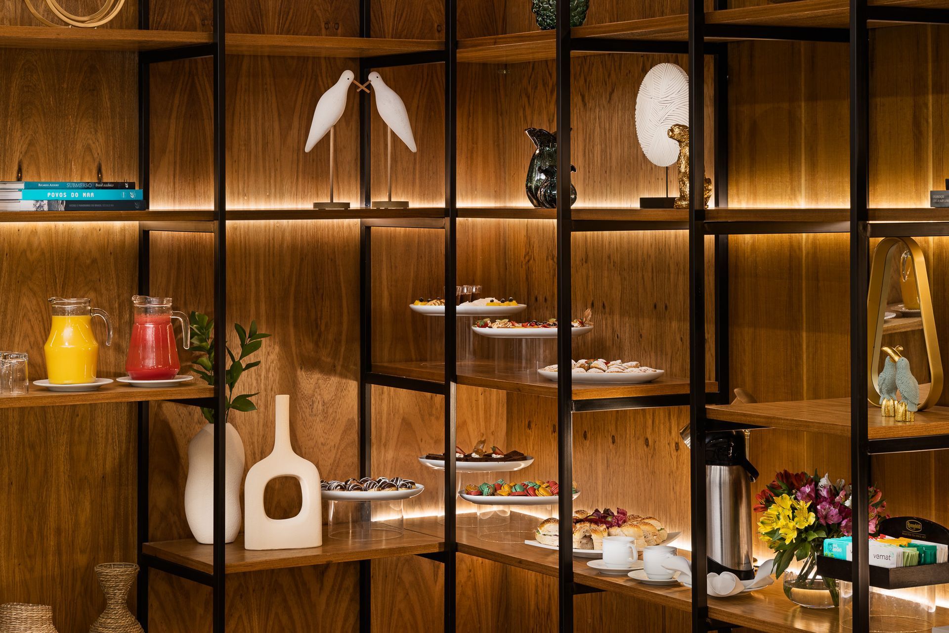 Estante com diversos itens alimentícios e de decoração: doces, bebidas, esculturas e flores. Othon