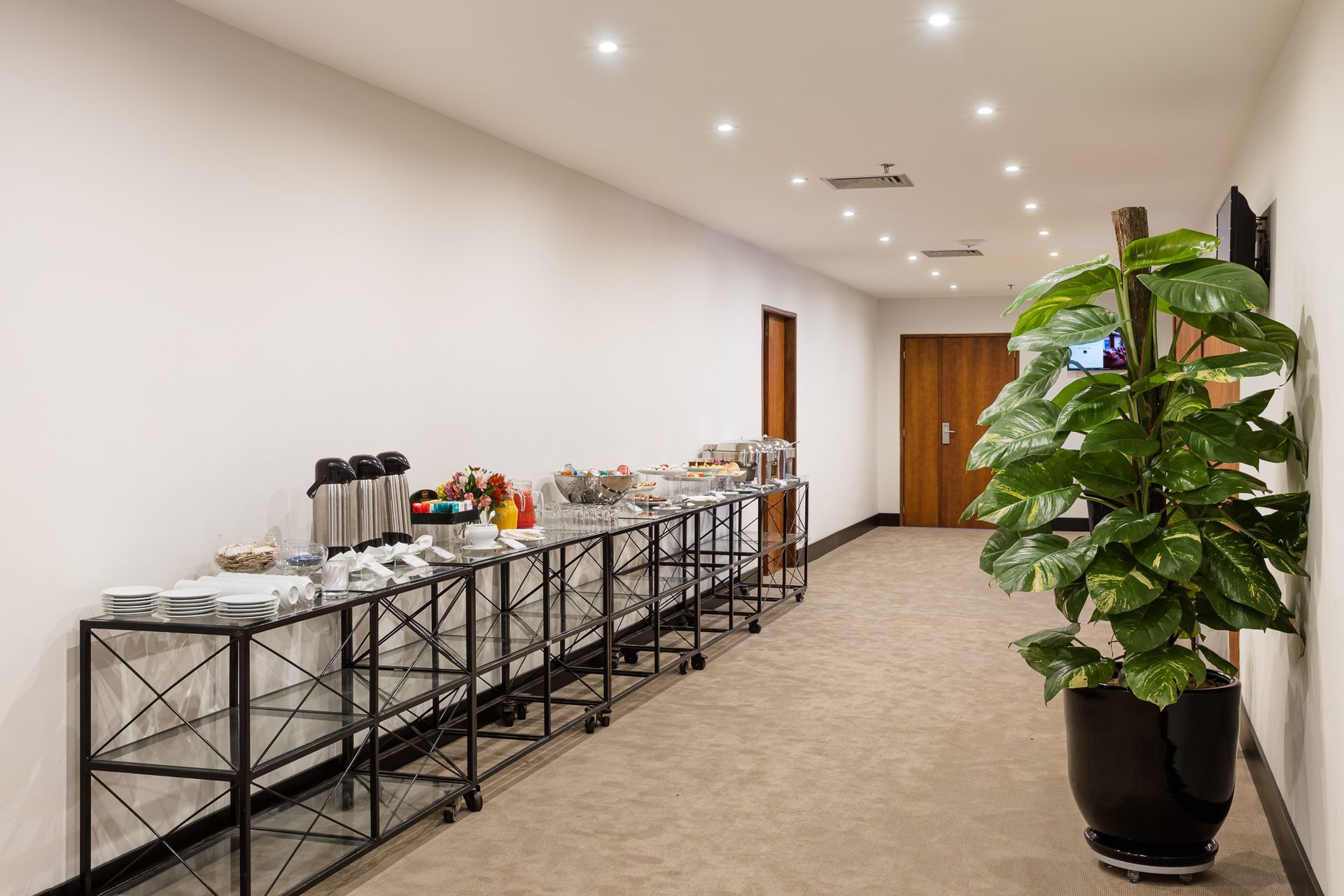 Corredor com mesas de bufê e um grande vaso de plantas. Othon
