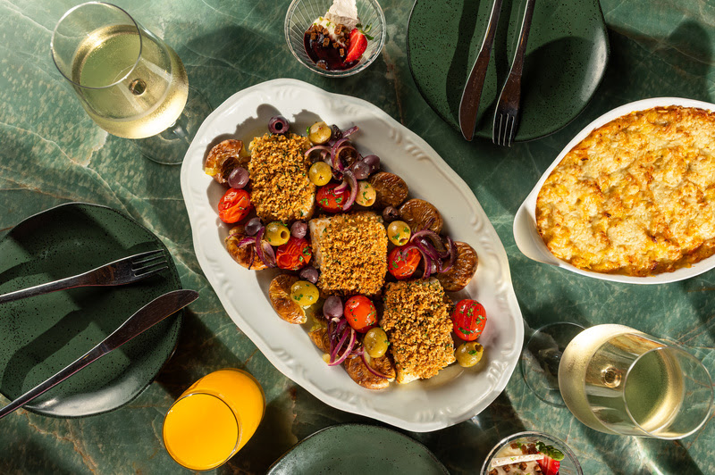 Bacalhau assado com batatas, ovos e azeitonas servido em travessa, acompanhado de vinho e acompanhamentos, em mesa posta elegante