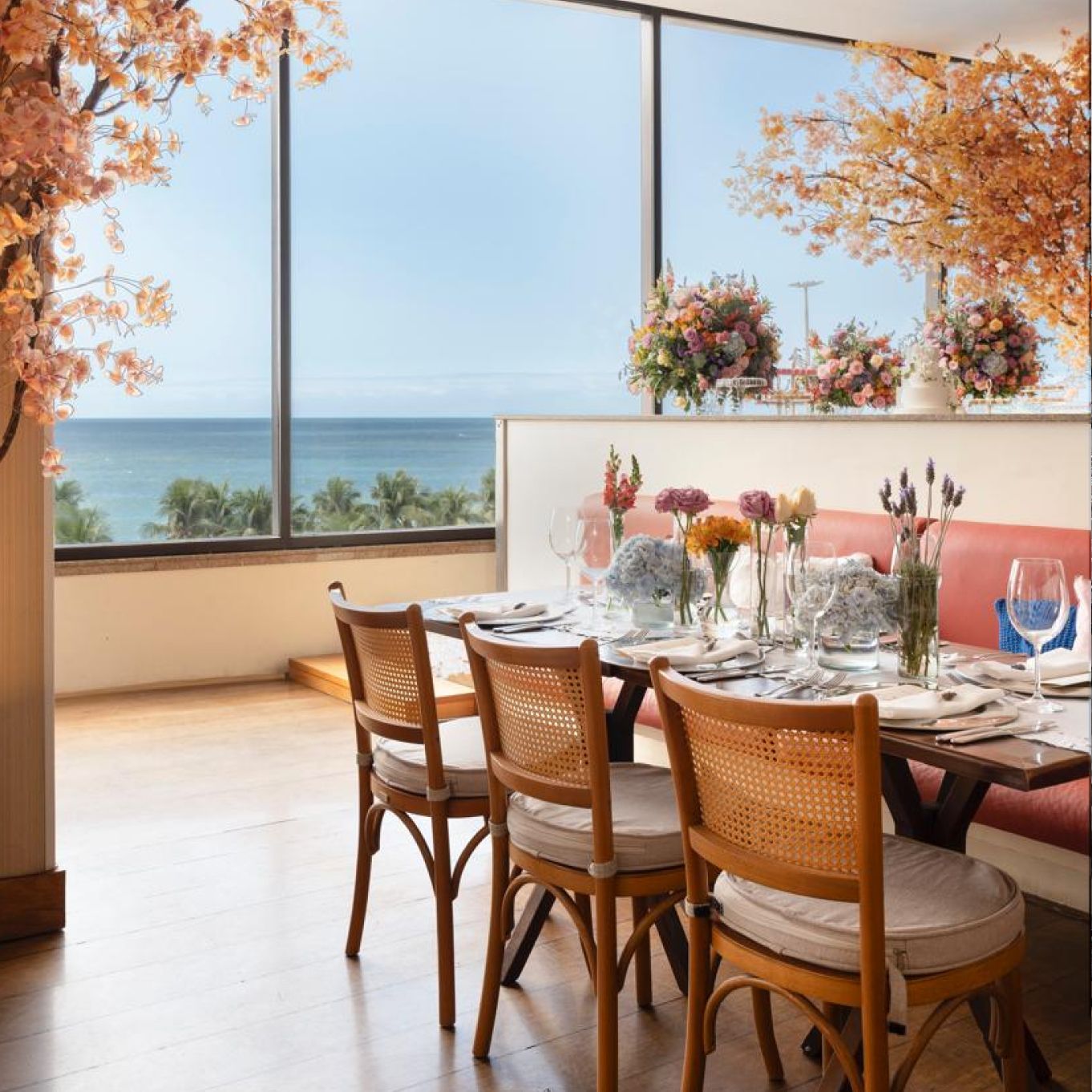 Sala de jantar com mesa posta para refeição, vista para o mar, decorada com flores e galhos de árvores de cores claras. Othon