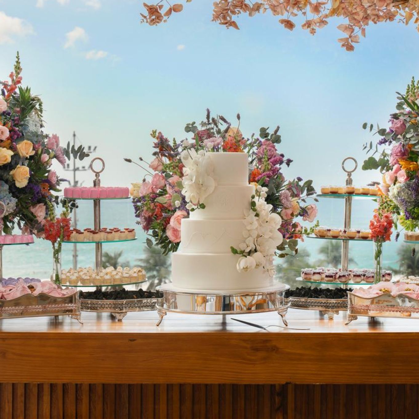 Bolo de casamento e sobremesas em uma mesa com vista para o mar e decoração floral. Othon