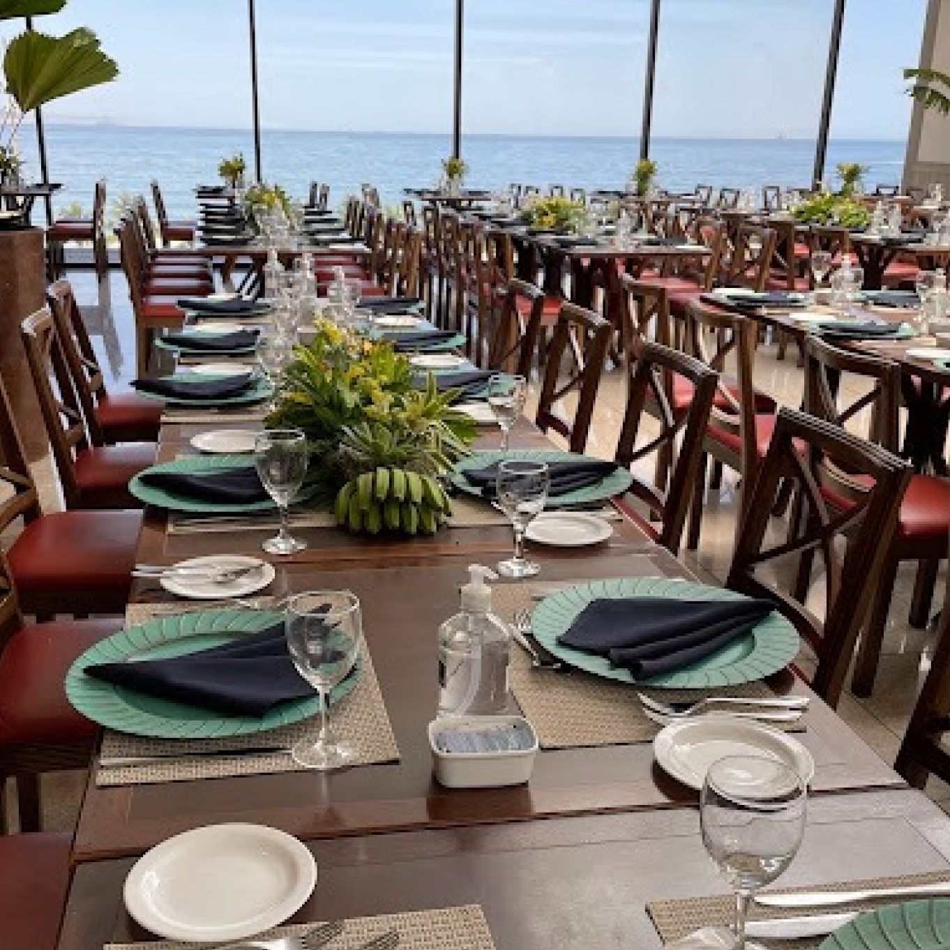 Mesas de restaurante preparadas para uma refeição, com vista para o mar. As mesas têm jogos americanos, pratos, guardanapos e arranjos florais. Othon