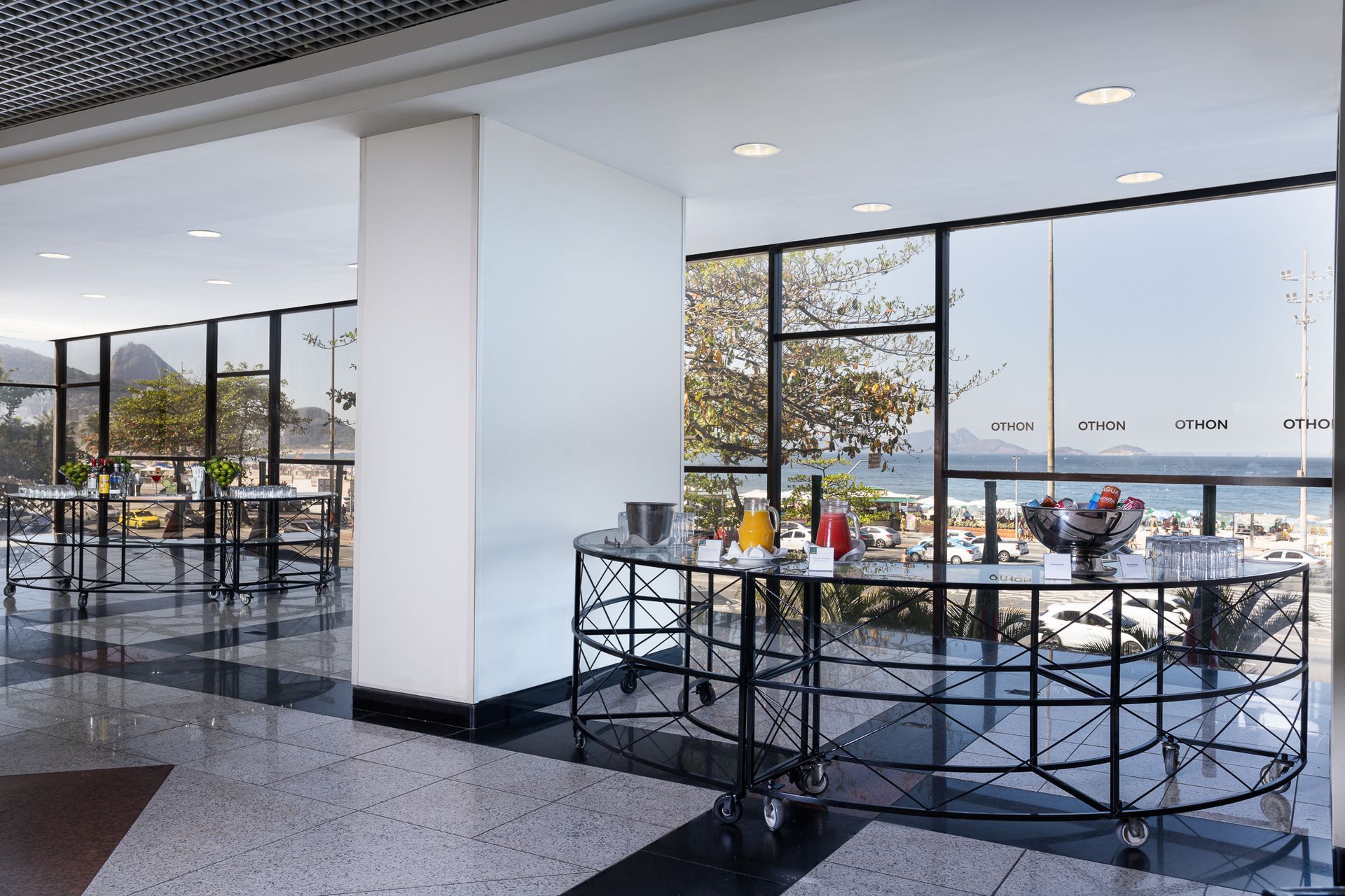 Área de refeições do hotel com vista para a praia. Mesas, carrinho de bufê com sucos e piso de cerâmica. Othon