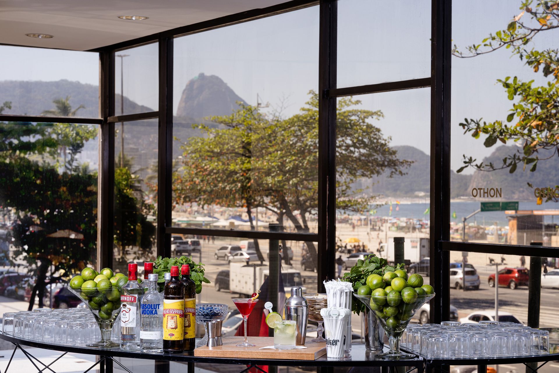 Bar com bebidas e copos, com vista para a praia e a montanha. Othon