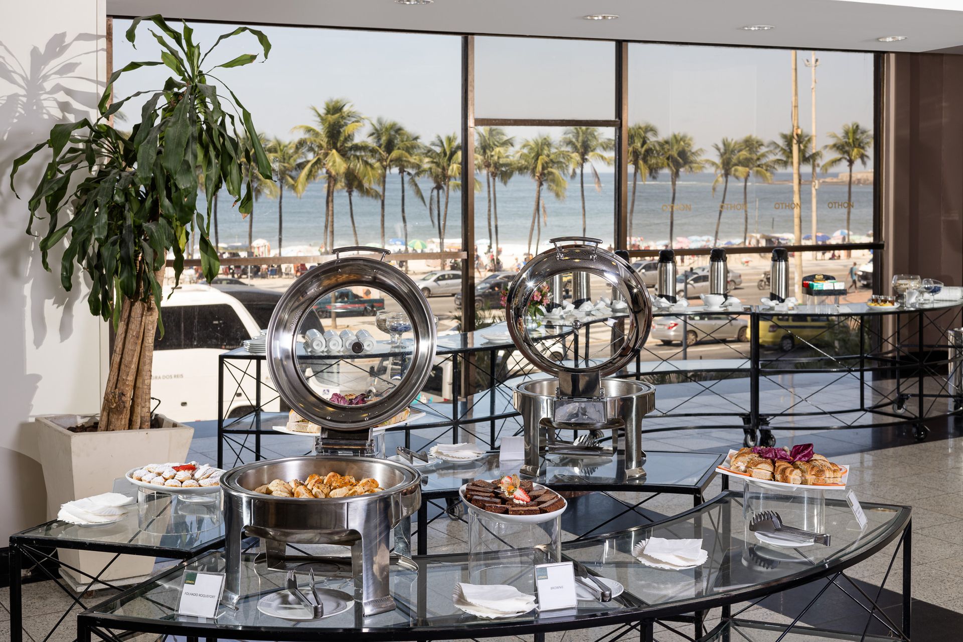 Mesa de bufê com comida em ambiente interno; vista da praia ao fundo. Othon