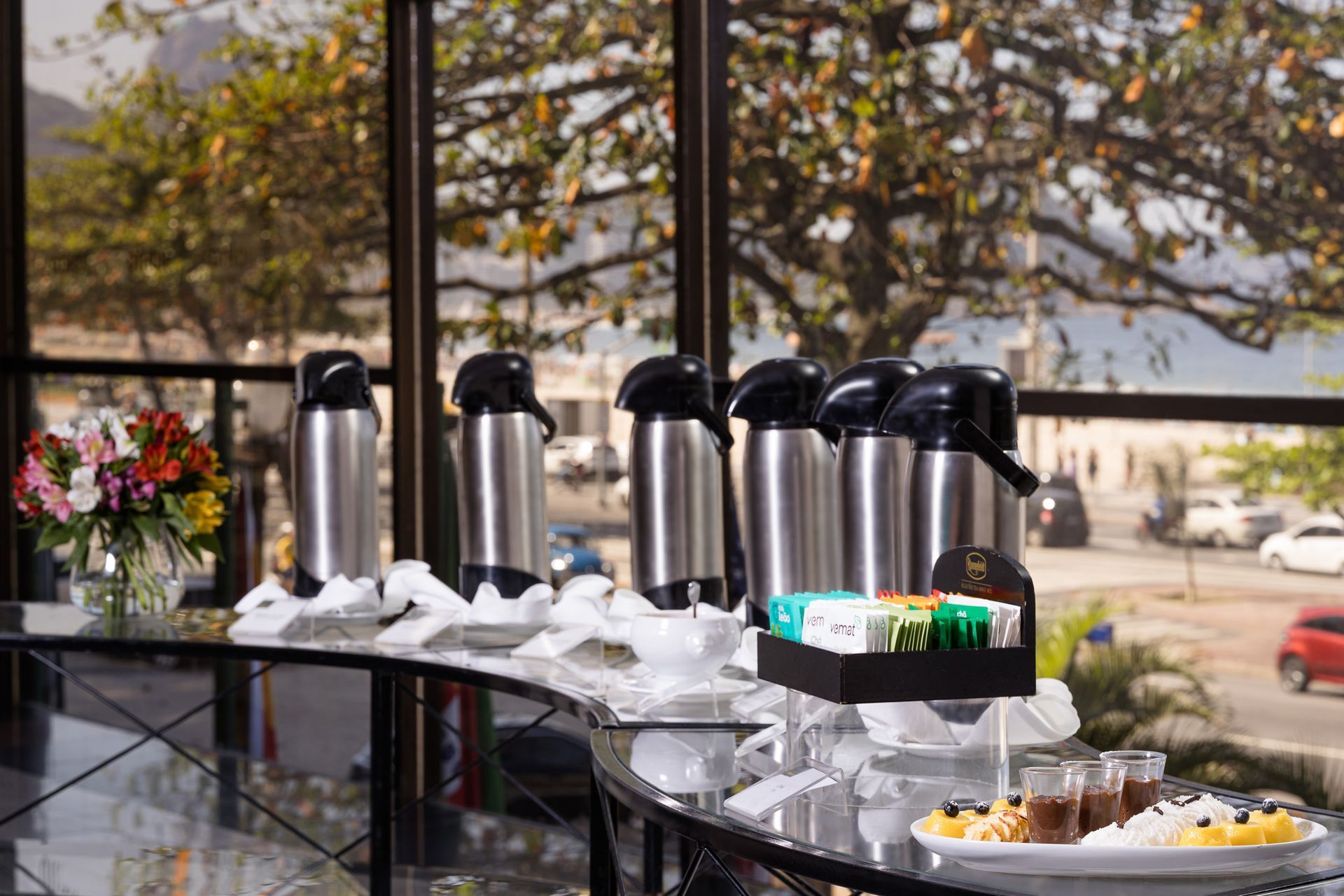 Estação de café com garrafas térmicas, chá e doces em um balcão perto de uma janela com vista para o exterior. Othon