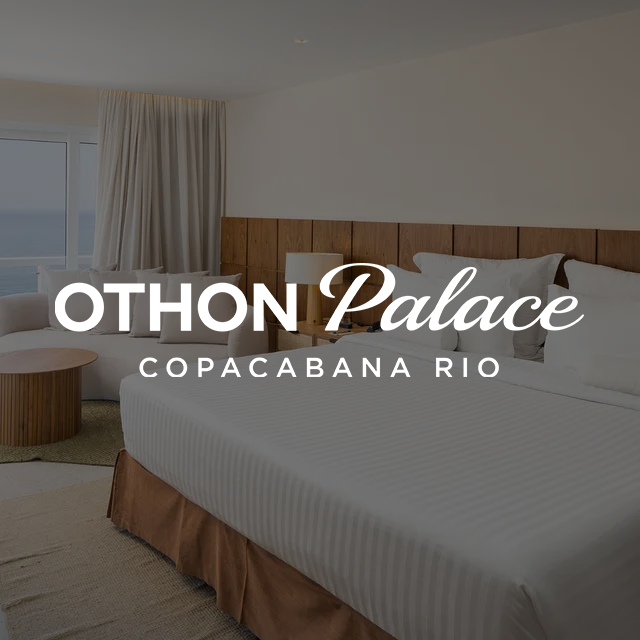 Quarto com cama, sofá, mesa de cabeceira e vista para o mar. Tons claros e neutros.