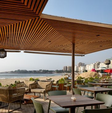 Restaurante ao ar livre com vista para a praia. Teto de madeira, mesas, cadeiras e vista para o oceano e edifícios.
