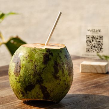 Bebida de coco com canudo sobre uma mesa de madeira, com um código QR ao fundo.