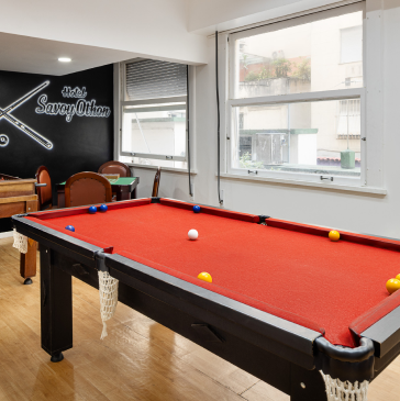 Mesa de bilhar com feltro vermelho em uma sala de jogos de hotel; placa do Hotel Savoy Urban.