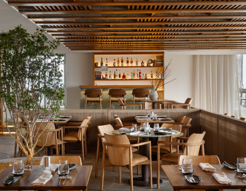 Interior de restaurante com mesas e cadeiras de madeira, bar e prateleiras de bebidas.