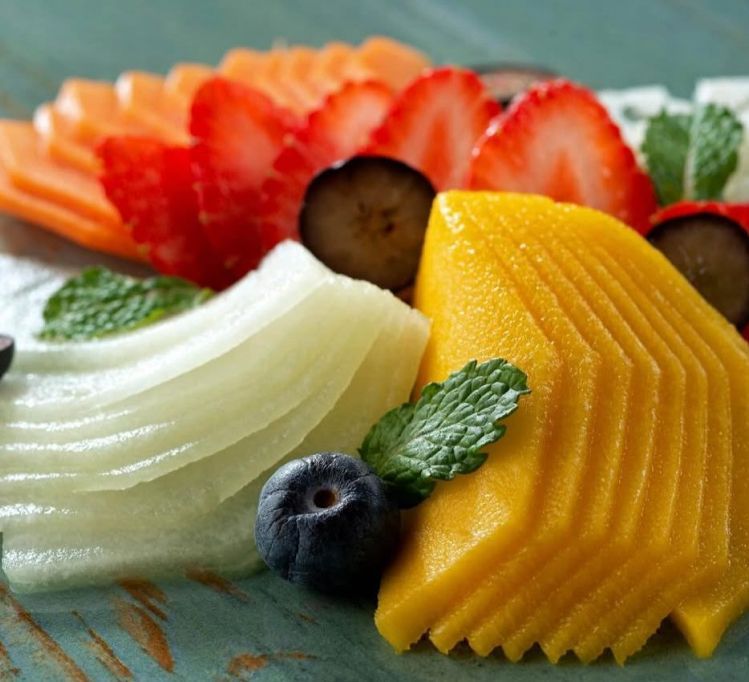 Prato de frutas fatiadas: melão, morangos, manga, mirtilos, hortelã, sobre madeira azul.