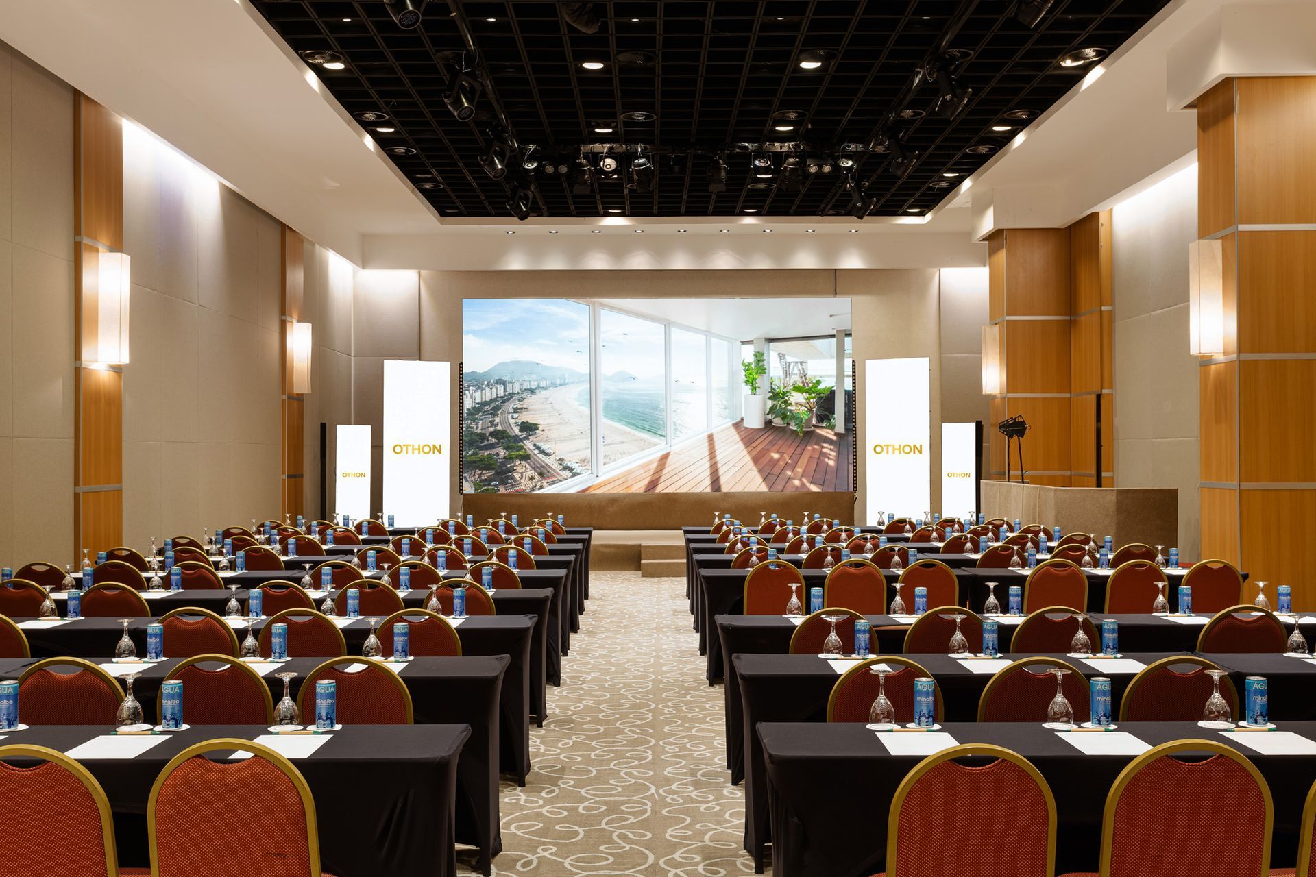 Sala de conferências com fileiras de mesas, uma tela grande exibindo uma sala e cadeiras vermelhas. Othon