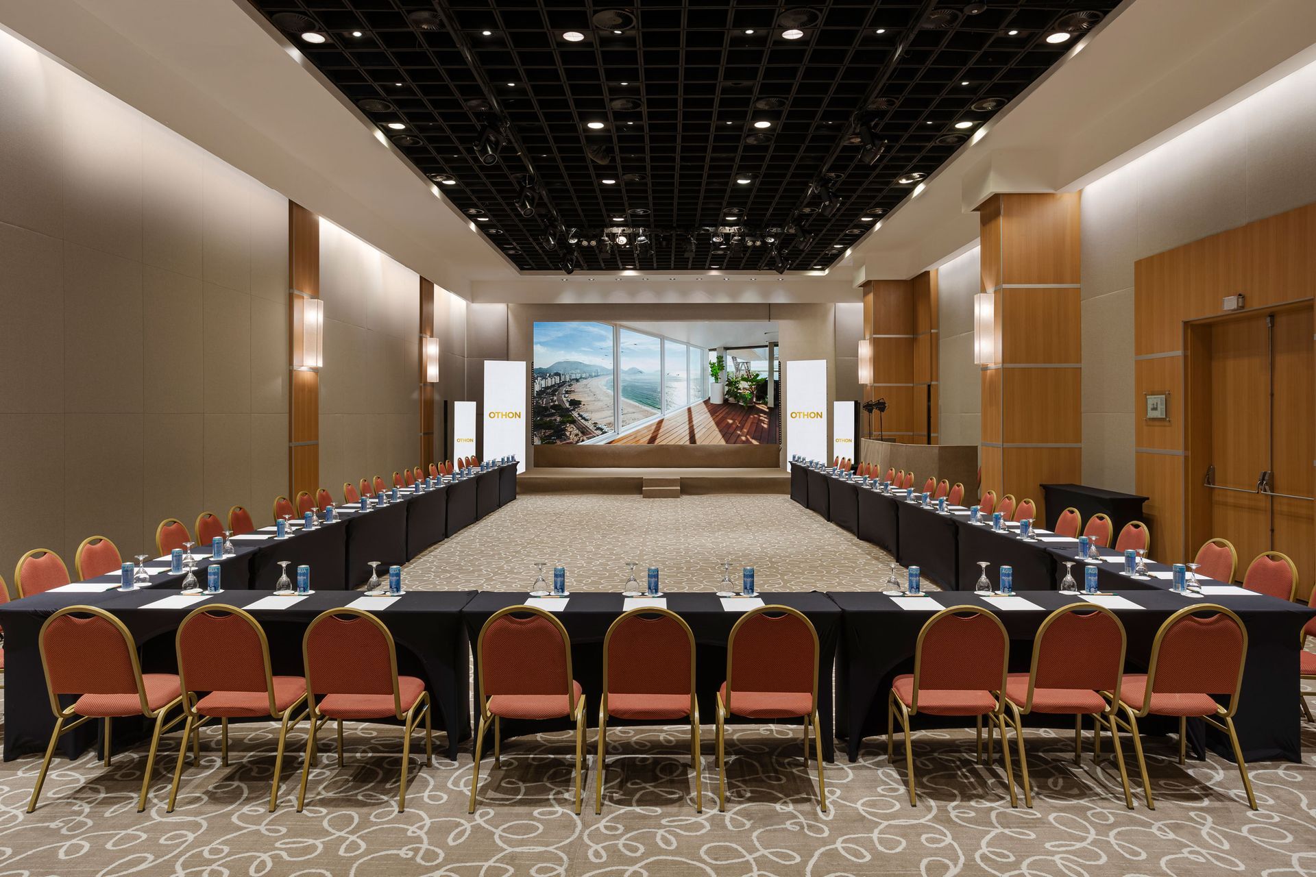 Sala de conferências com mesas dispostas em formato de U, de frente para uma tela, cadeiras e painéis de madeira. Othon