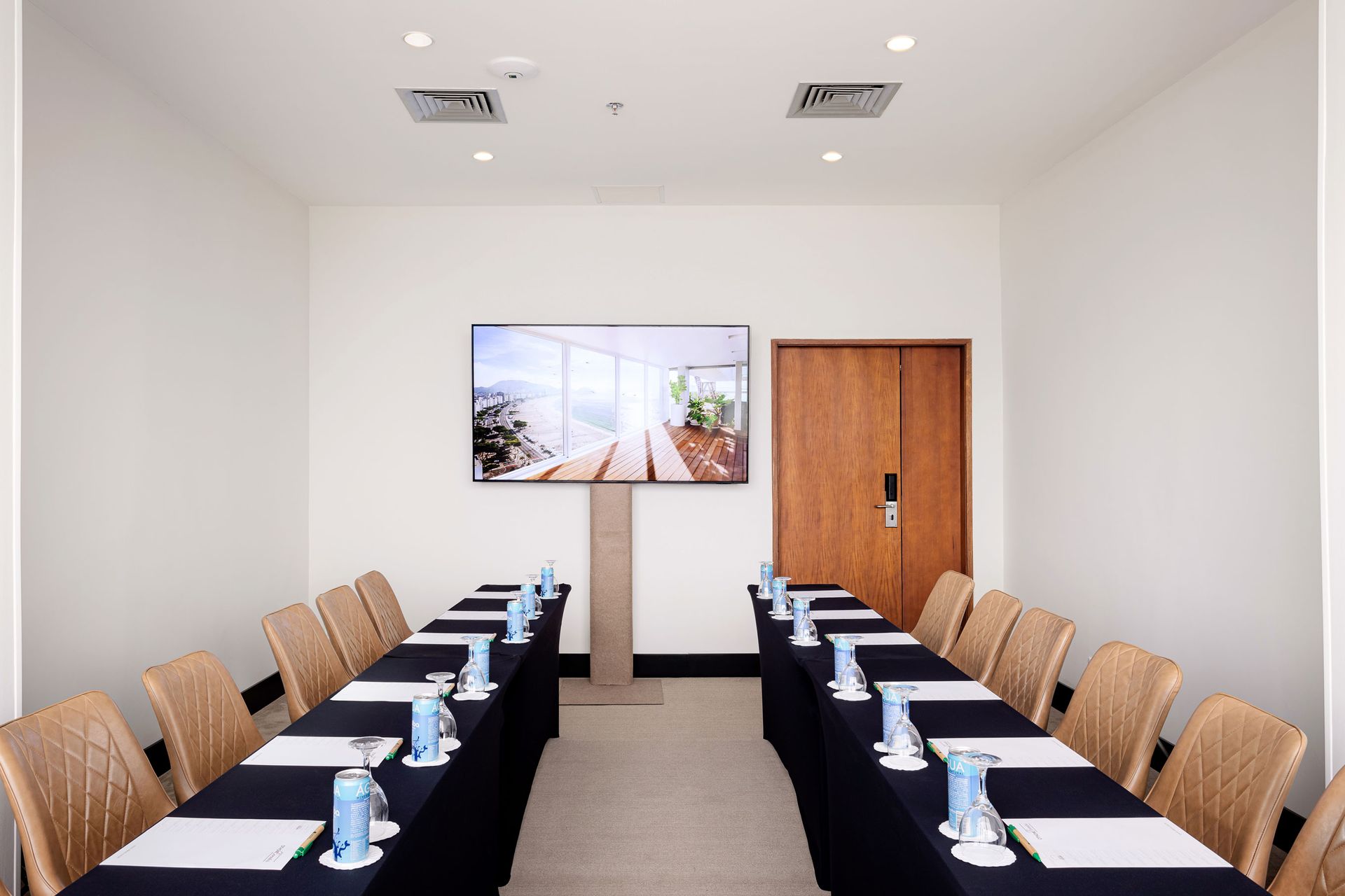 Sala de conferências com mesa longa, cadeiras, TV e porta fechada. Othon