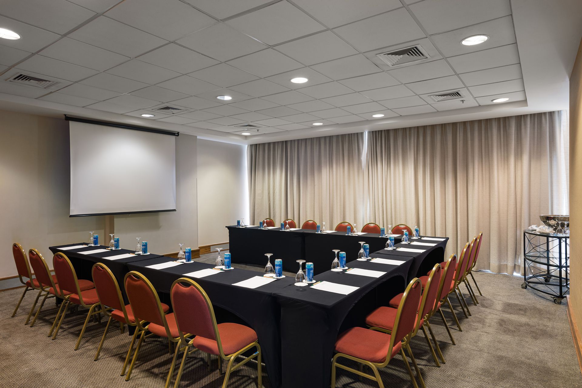 Sala de conferências com mesas em formato de U, cadeiras vermelhas, tela de projetor e cortinas bege. Othon