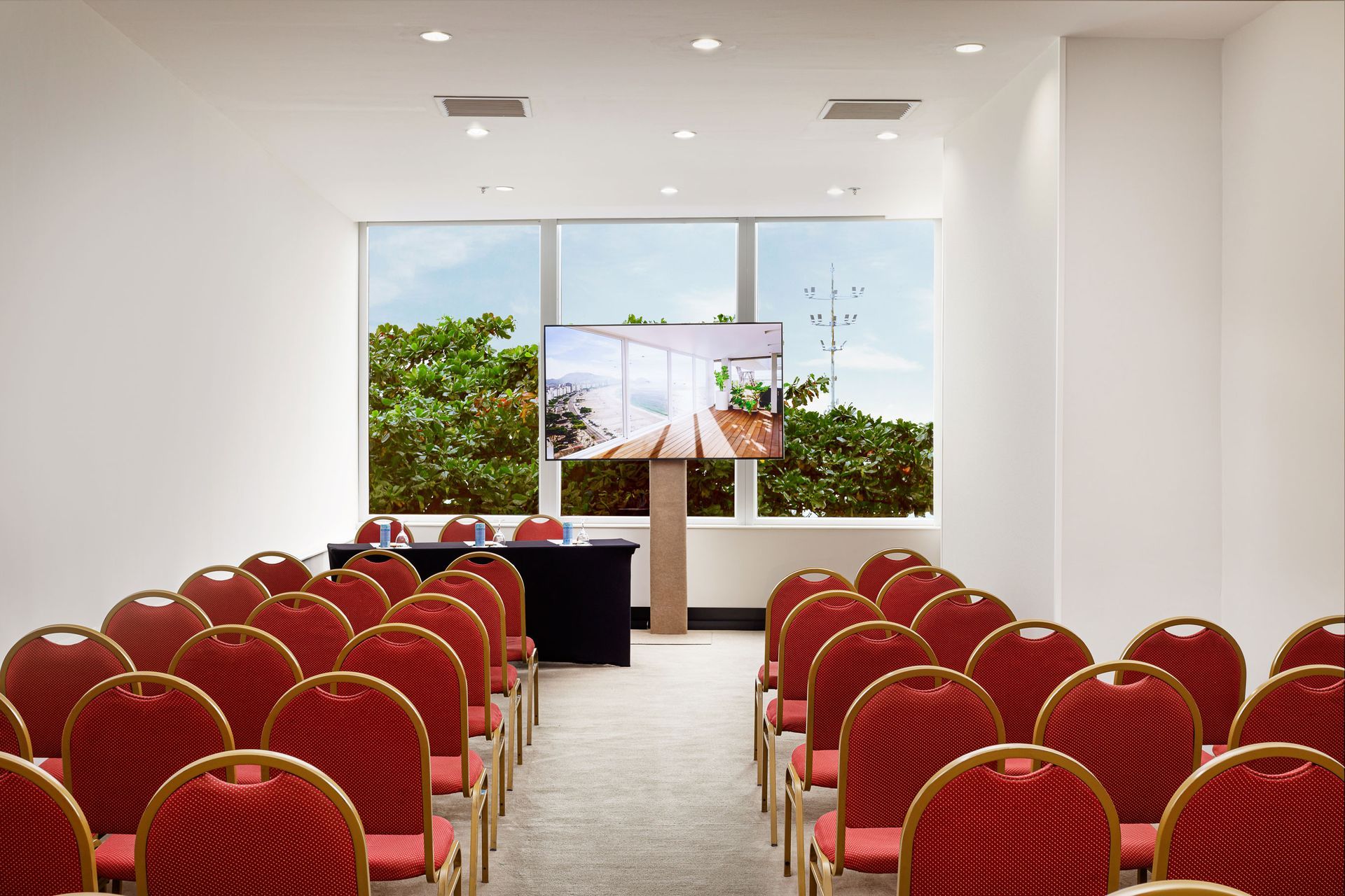 Sala de conferências com cadeiras vermelhas dispostas em fileiras, de frente para uma tela e uma janela. Othon