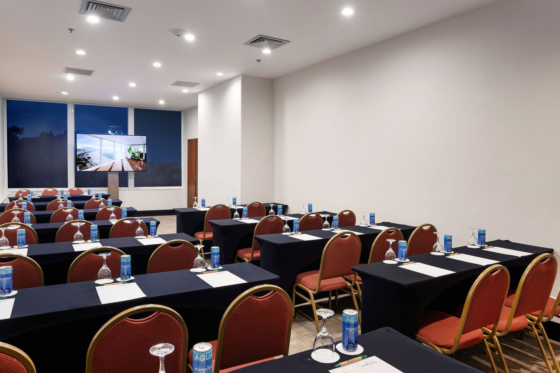 Sala de conferências com mesas, cadeiras e uma tela, preparada para uma apresentação. Othon