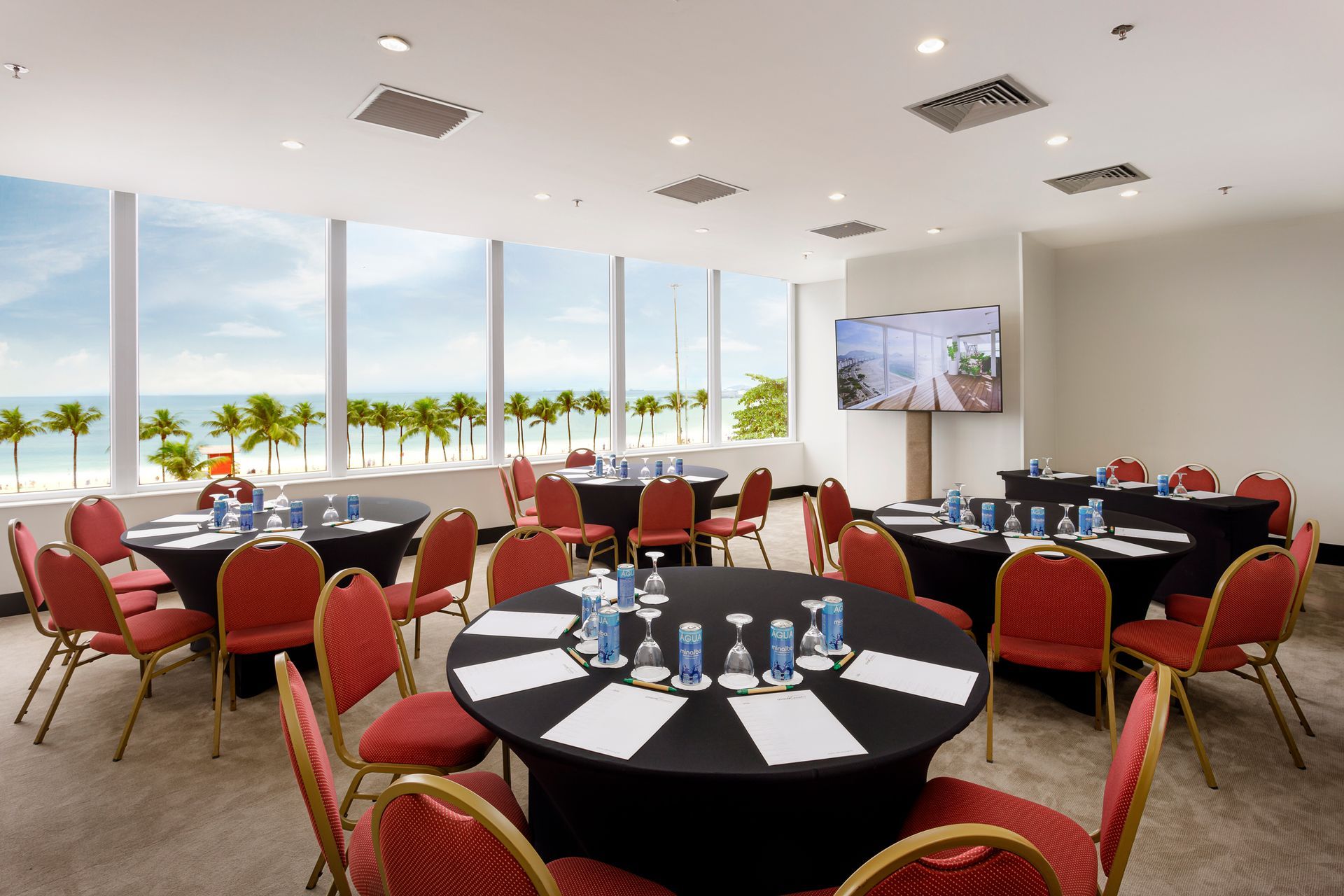 Sala de reuniões com mesas redondas, cadeiras vermelhas e vista para o mar através de grandes janelas. Othon