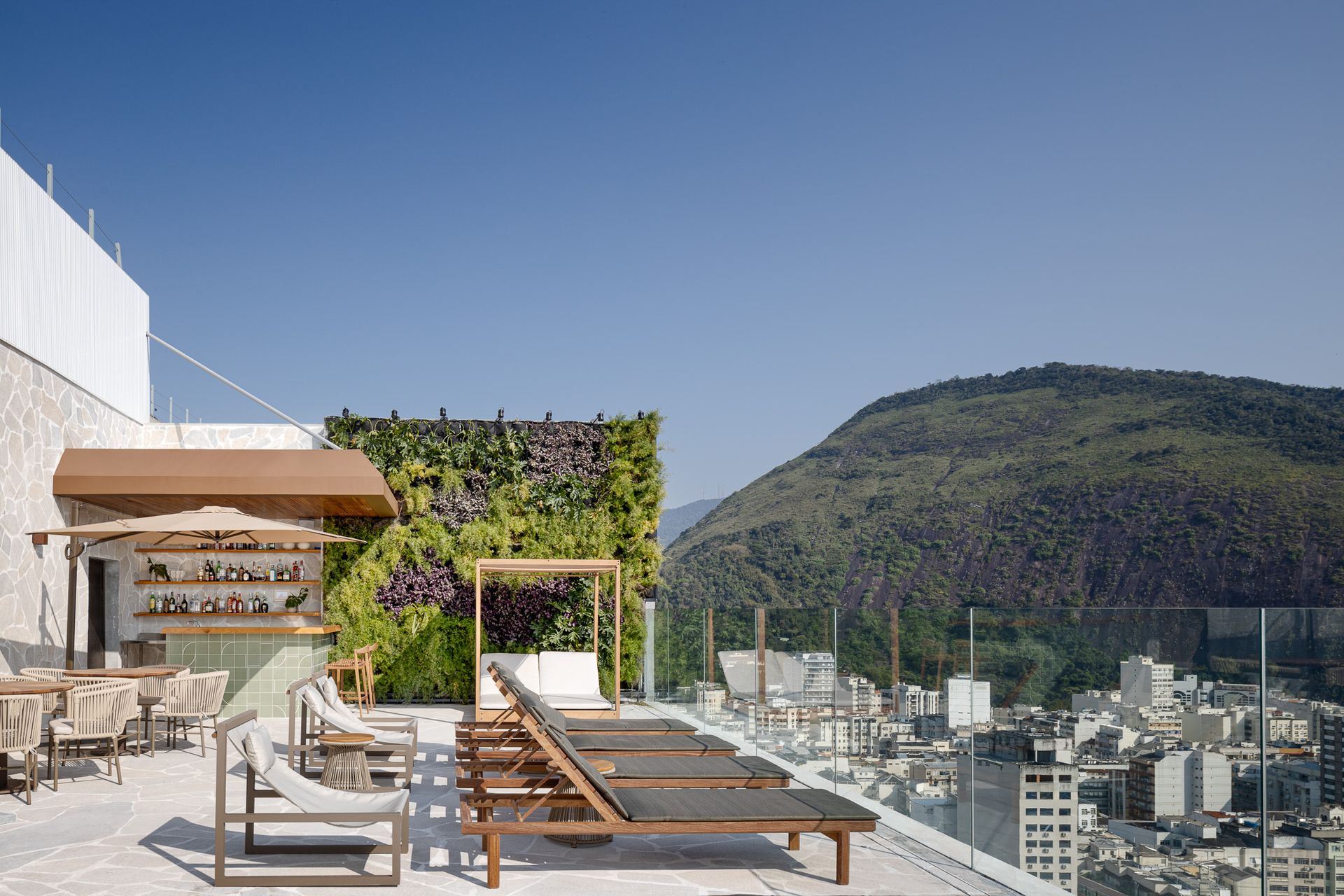 Terraço na cobertura com espreguiçadeiras, bar e parede verde com vista para a cidade e a montanha sob um céu azul.