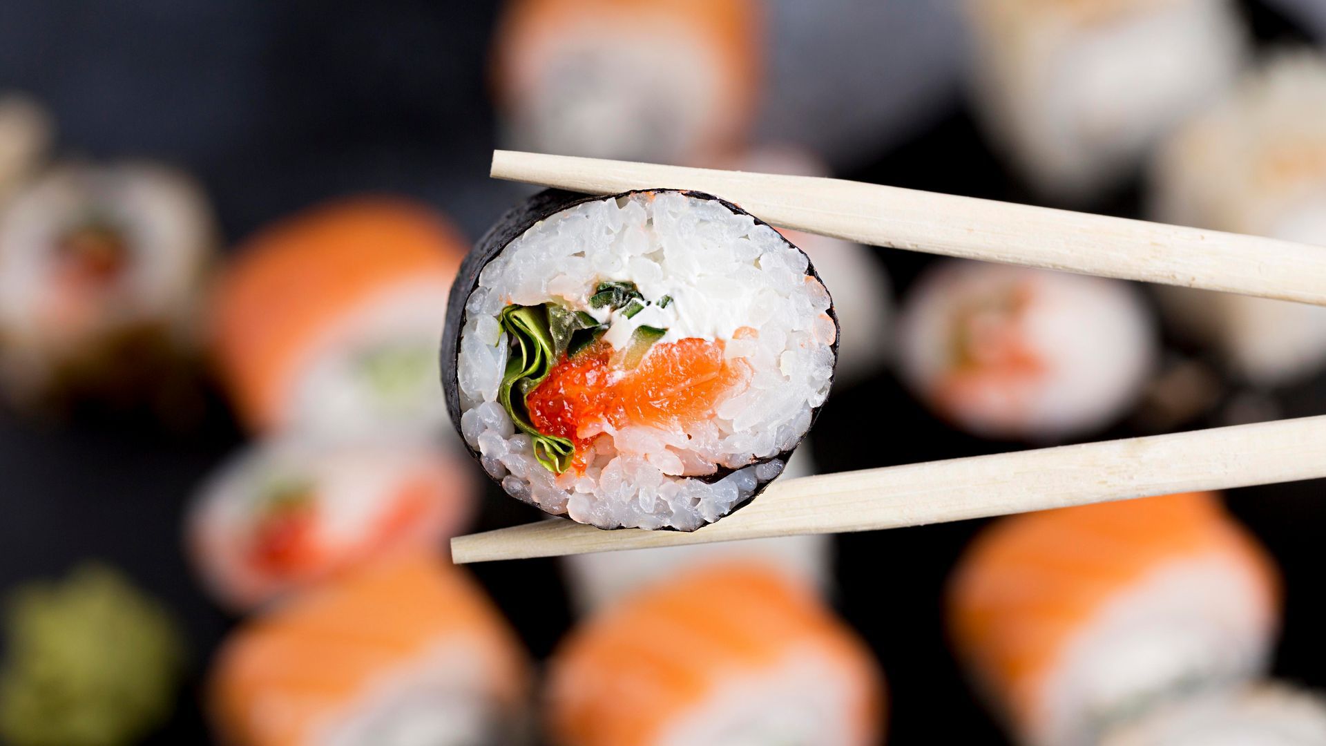 Rolo de sushi segurado por hashis, close-up com salmão, arroz e alga marinha. Outros rolos desfocados ao fundo.