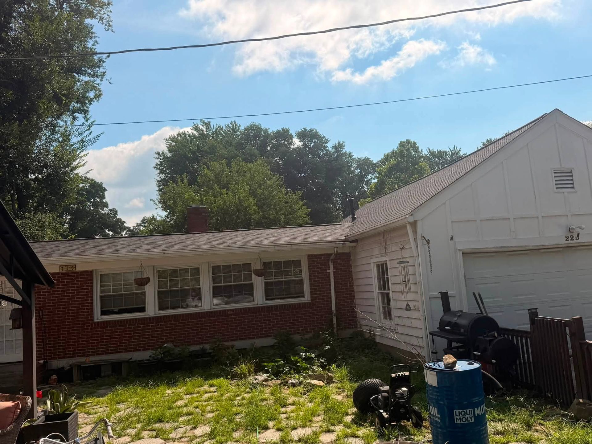 Small backyard with a red and white house, garage, trees, and a blue barrel under a bright sky