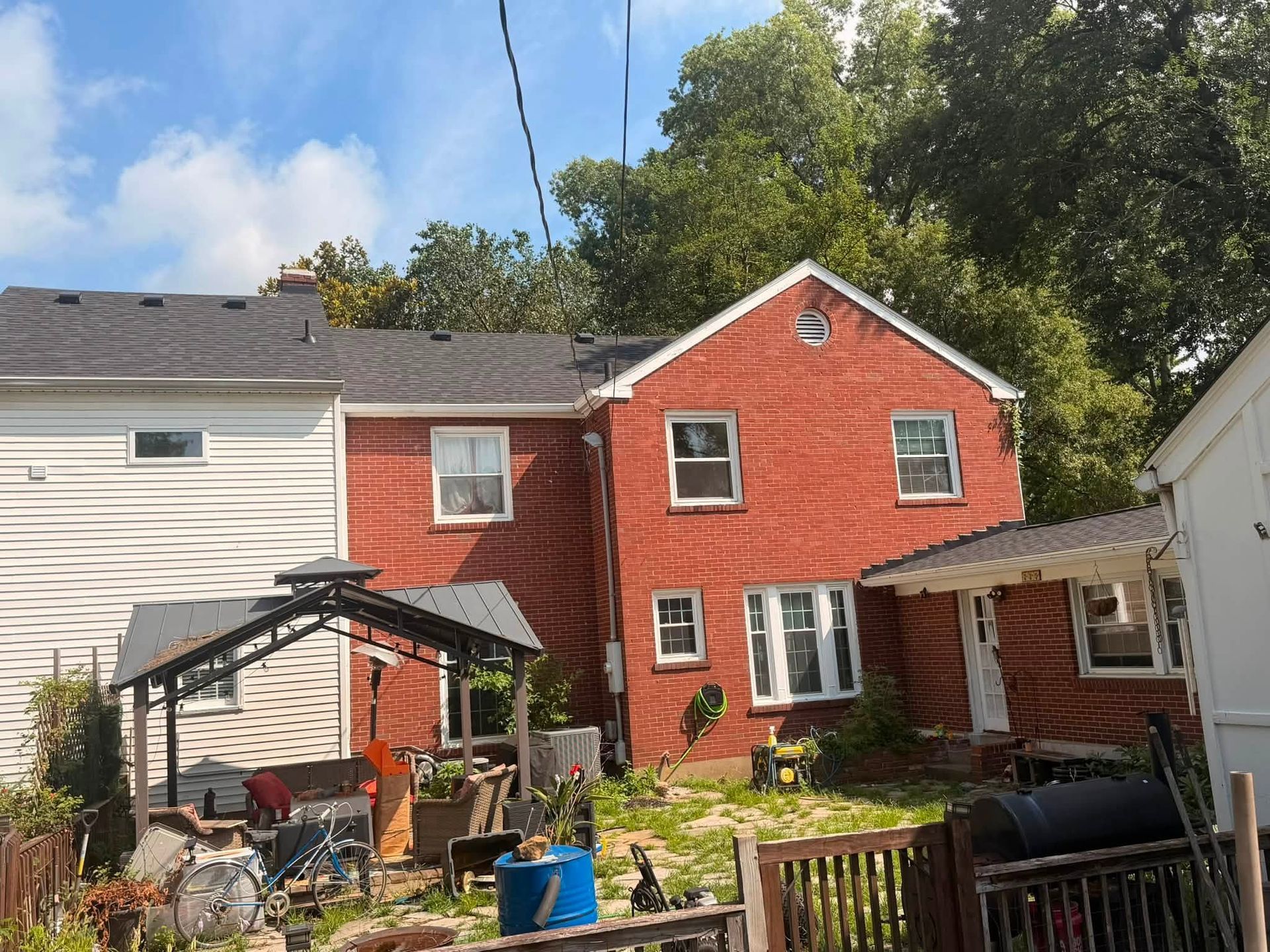 Red two-story house with backyard patio, grill, and scattered outdoor items under a blue sky