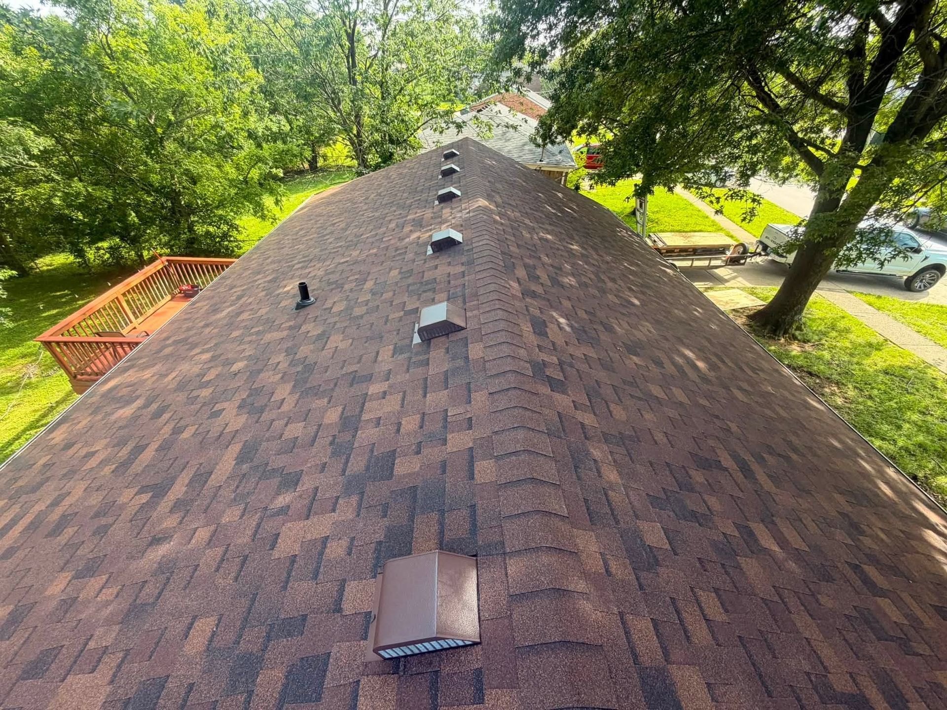 Aerial view of a shingled roof with several vents, surrounded by green trees and a driveway