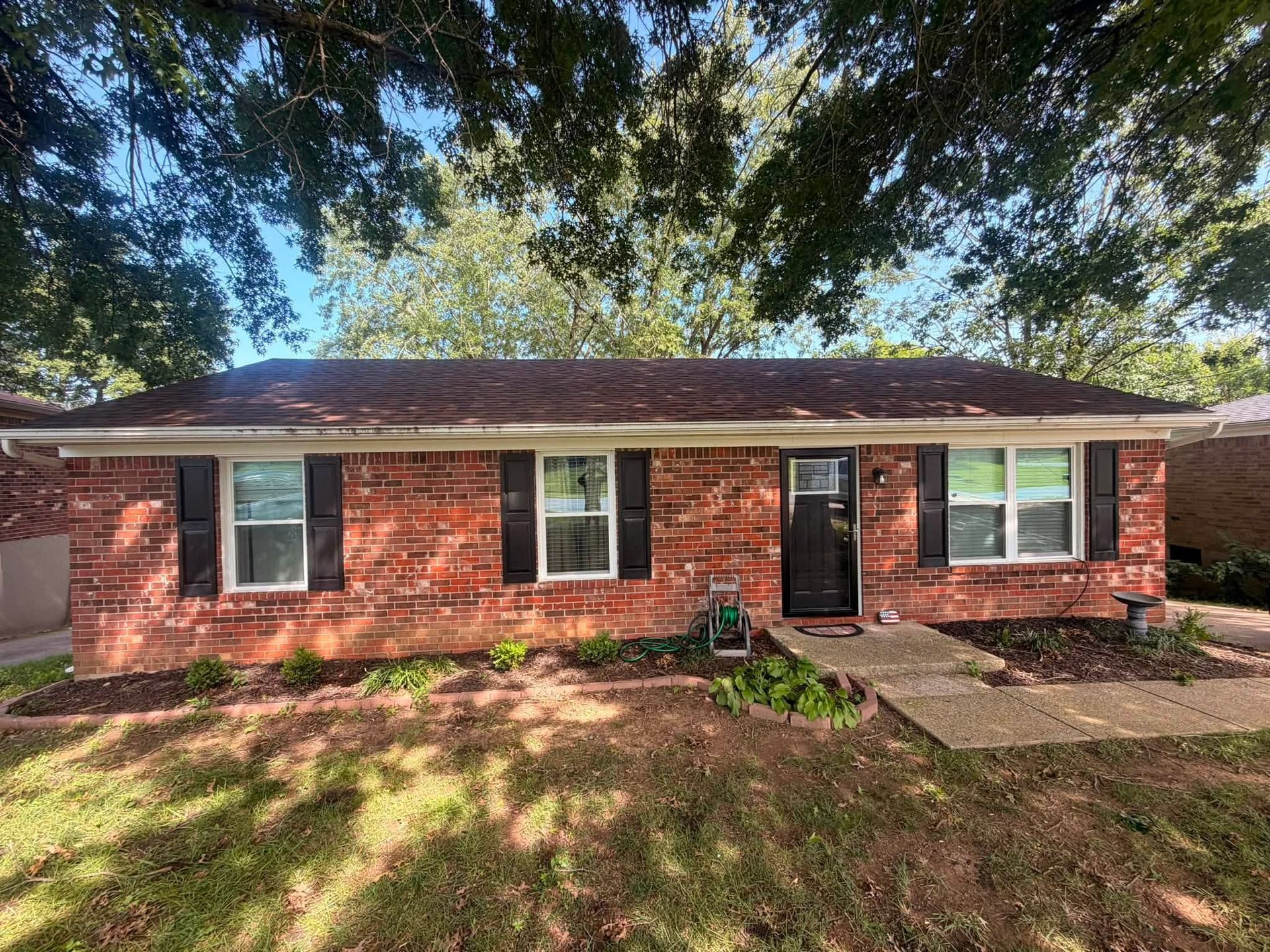 Single-story red brick house with front porch, trees overhead, and a grassy yard