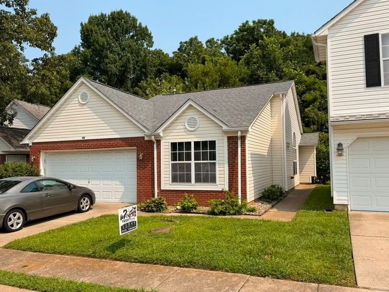 Small brick-and-beige suburban house with a garage, front lawn, and parked car in the driveway.