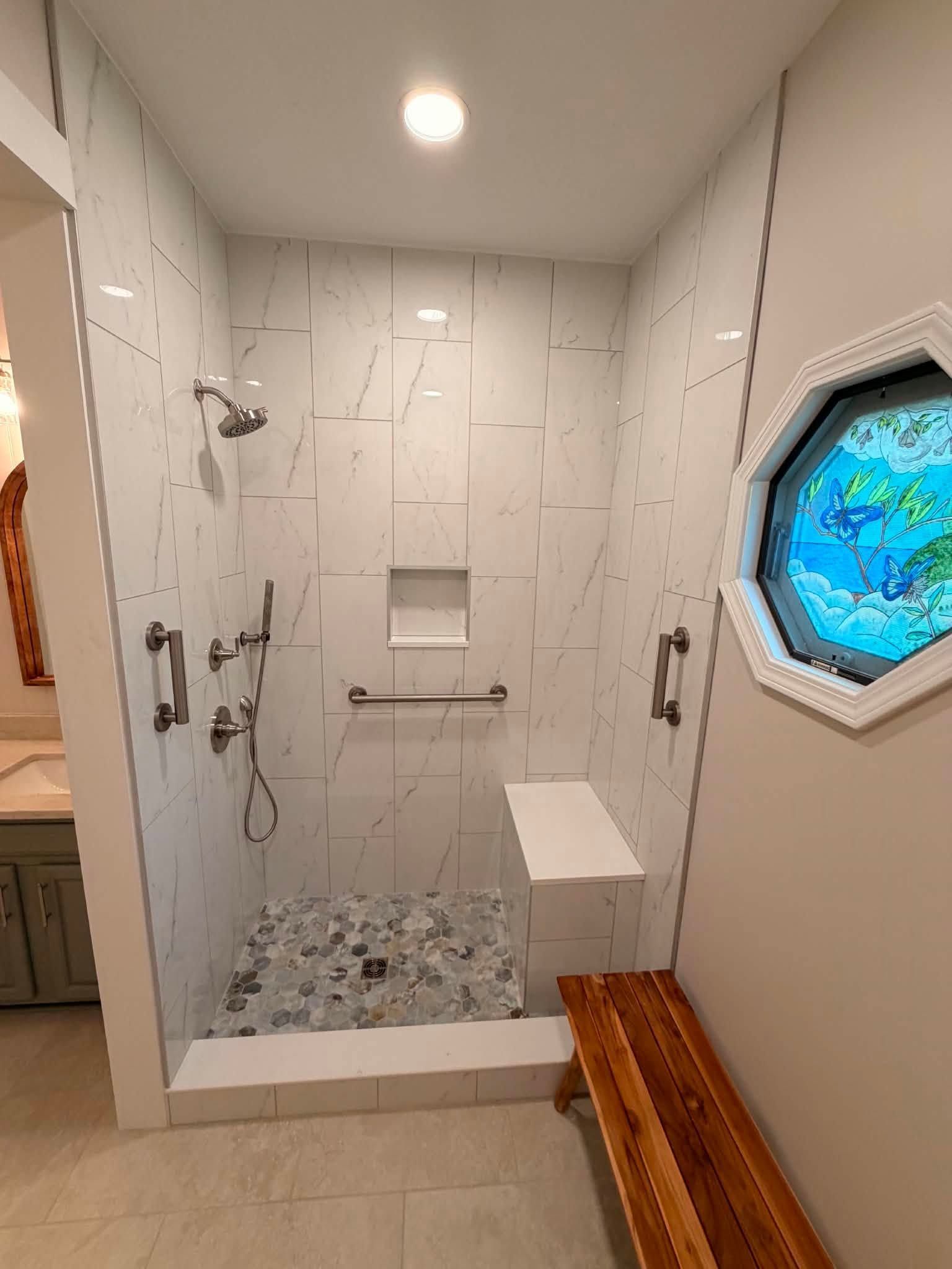 Accessible bathroom shower with white tile, grab bars, bench, and octagonal window.
