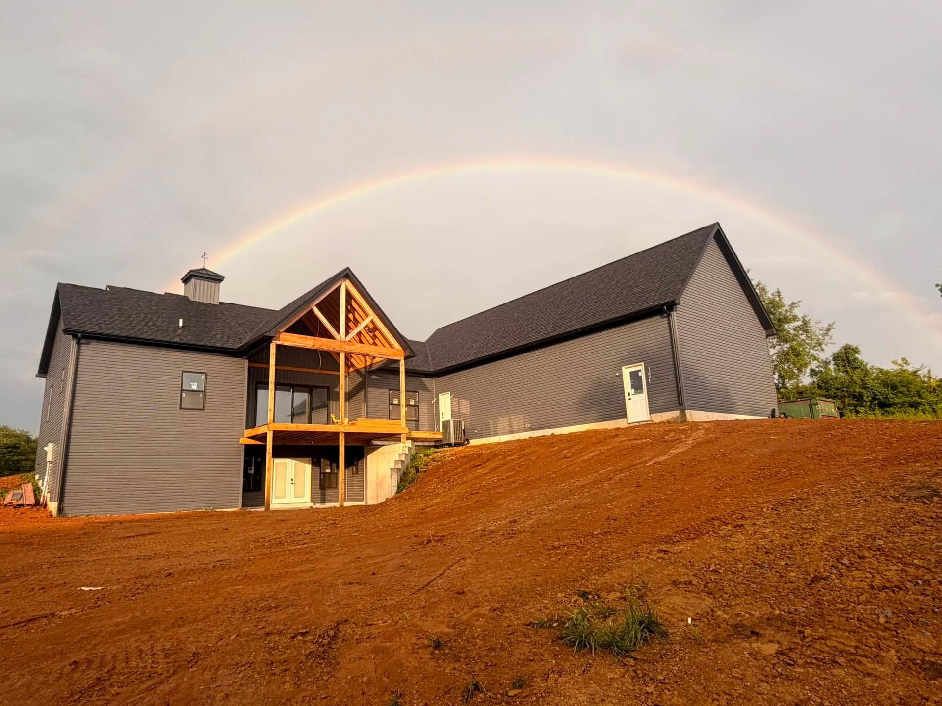 Modern gray house on a hillside with a wooden porch and a faint rainbow in the sky