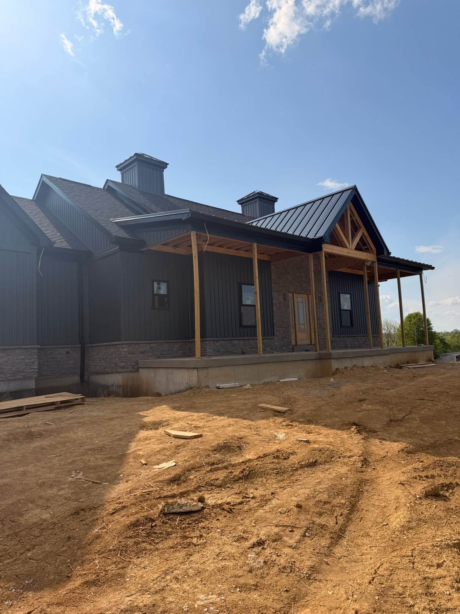 Partially built house with dark roof and wooden porch on a dirt lot under a blue sky