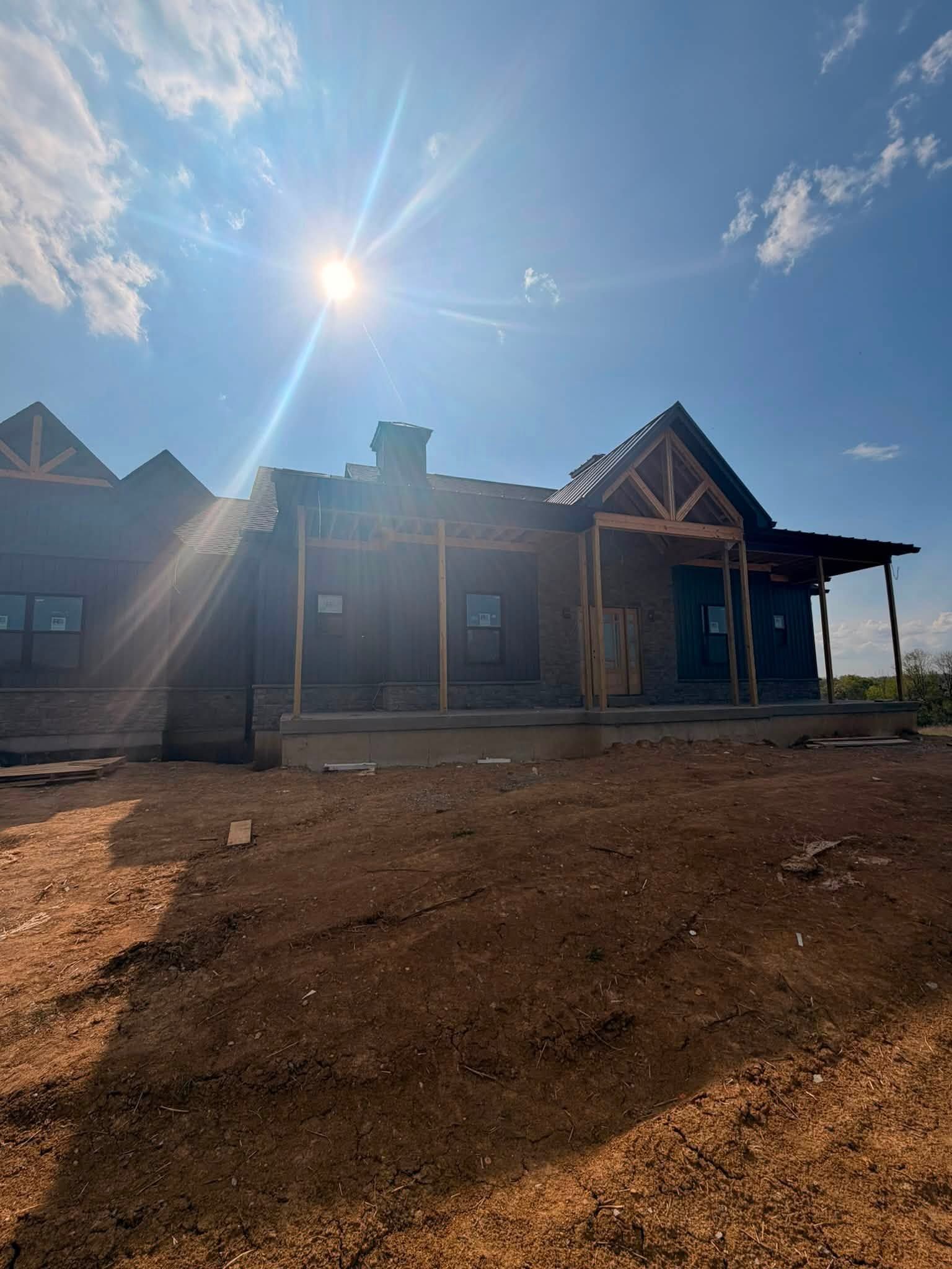 Sunlit unfinished house with porch and exposed dirt yard under a blue sky