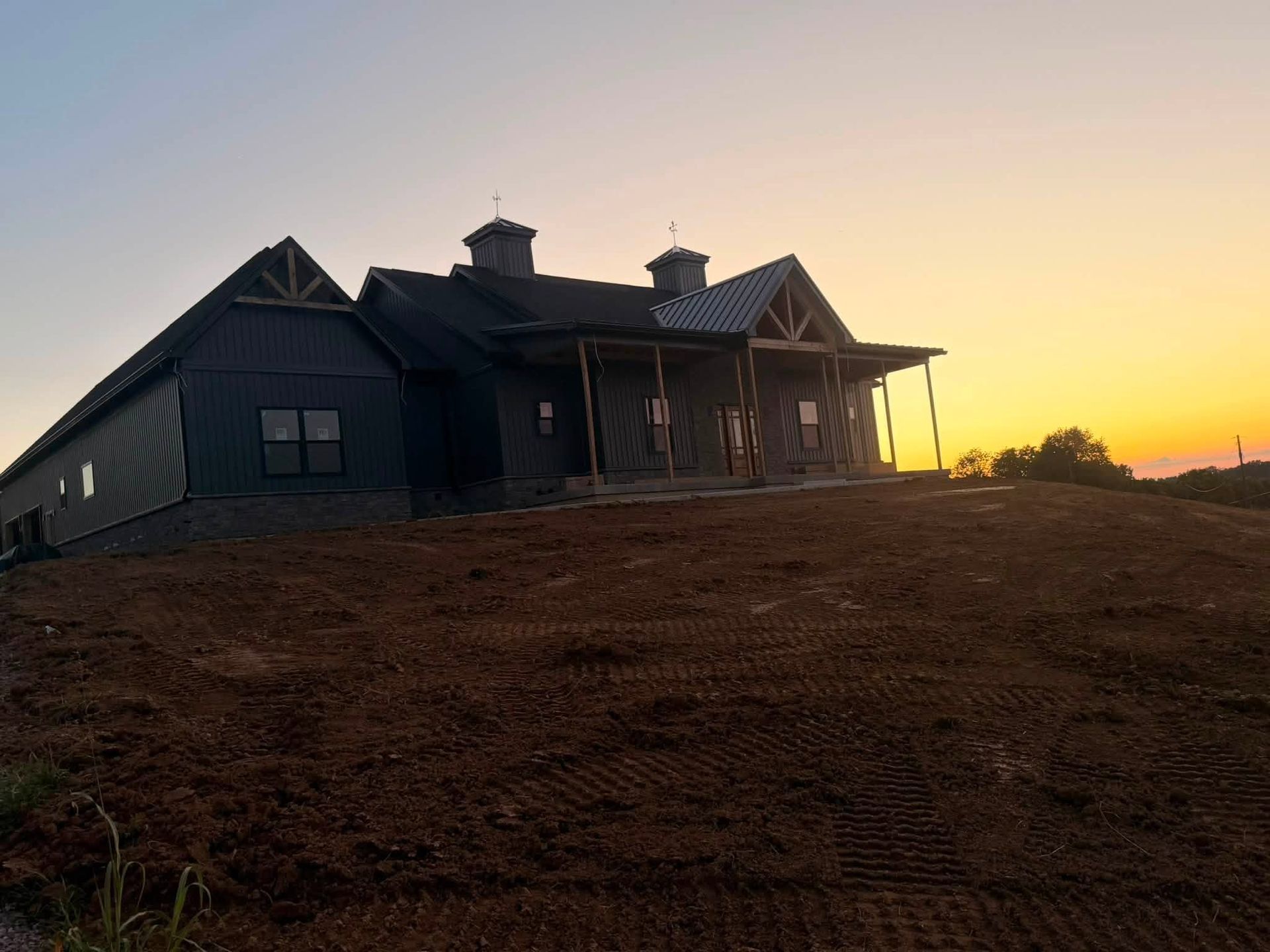 House under construction at sunset on a dirt hillside, silhouetted against a warm sky.