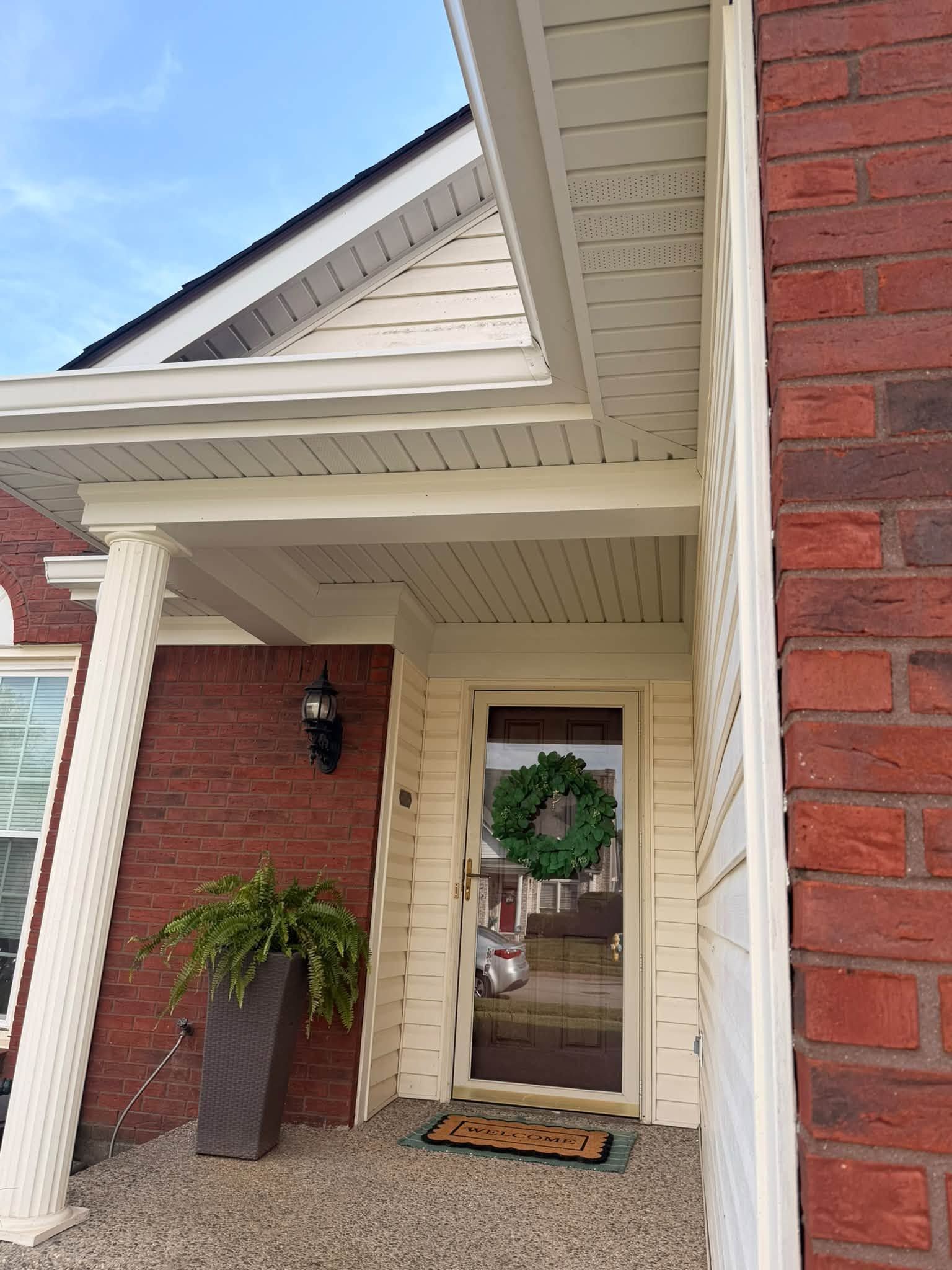 Front porch with red brick walls, white columns, wreath on the door, and a potted plant.
