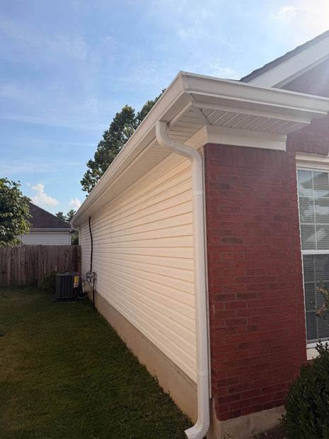 Side of a cream house with red brick corner, white trim, and a narrow grassy yard under a blue sky.