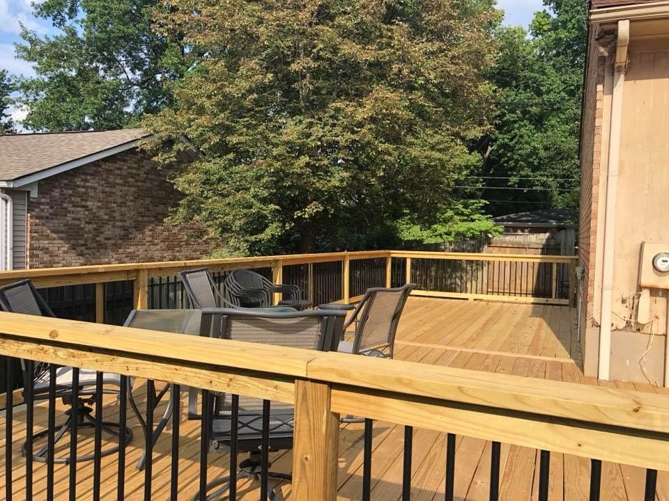 Wooden deck with patio table and chairs, surrounded by railings and trees in a sunny backyard